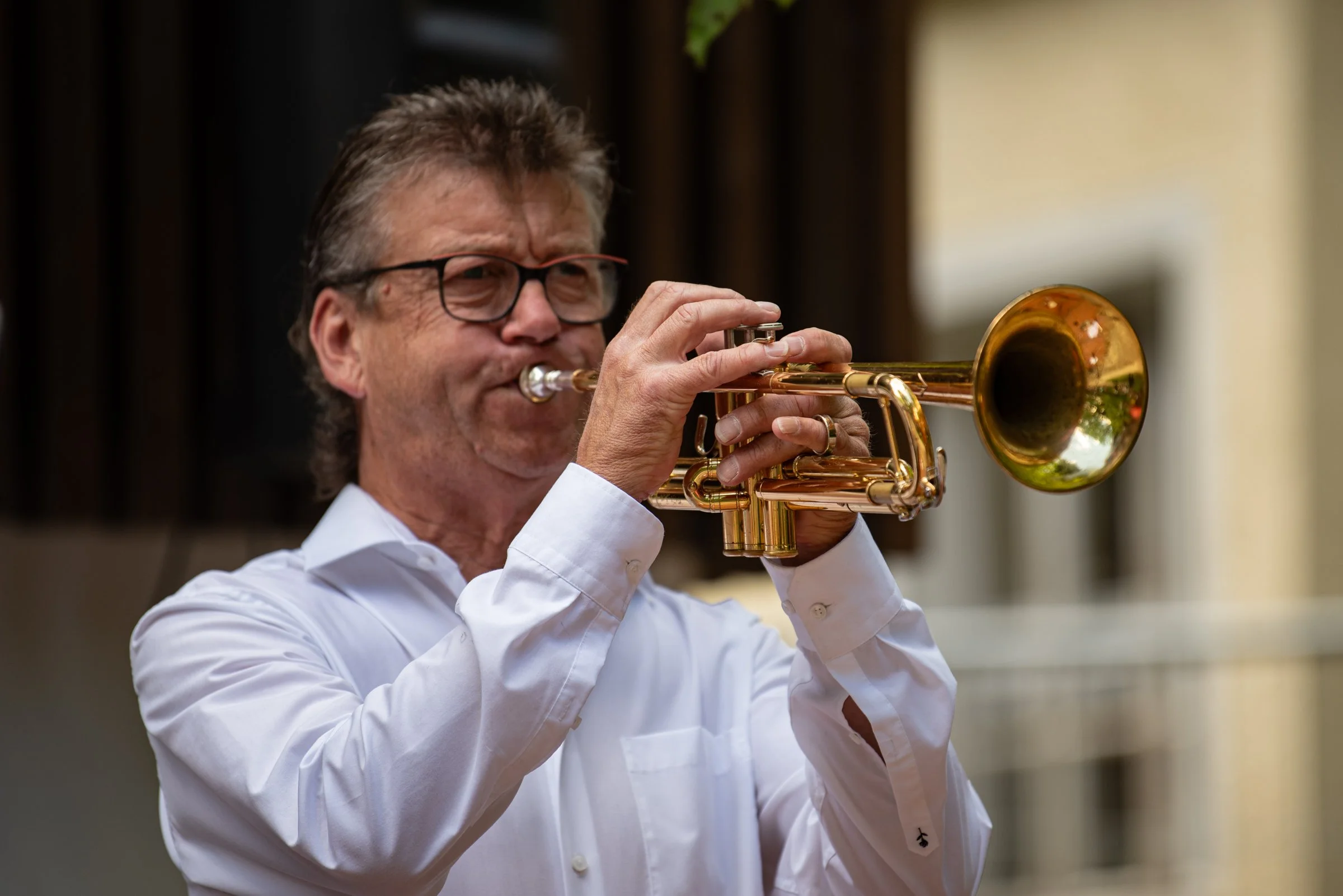 Ein Mann spielt Trompete im Freien, trägt eine weiße Hemd und trägt Brille.