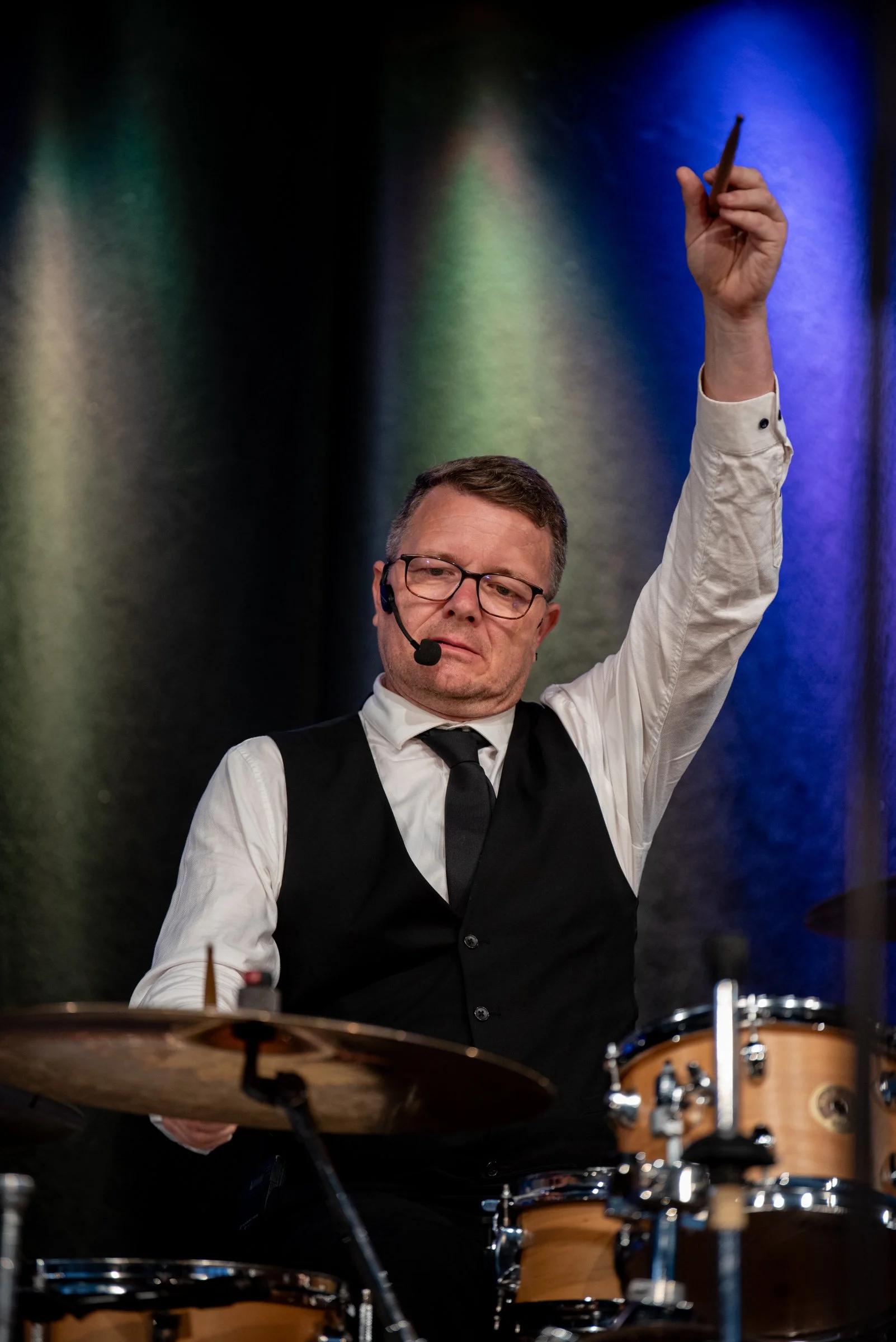 Ein Mann spielt Schlagzeug auf einer Bühne, trägt eine weiße Hemd, schwarzen Anzug und eine schwarze Krawatte, trägt ein Headset, hebt eine Hand mit einem Drumstick, im Hintergrund ist ein bunten Licht.