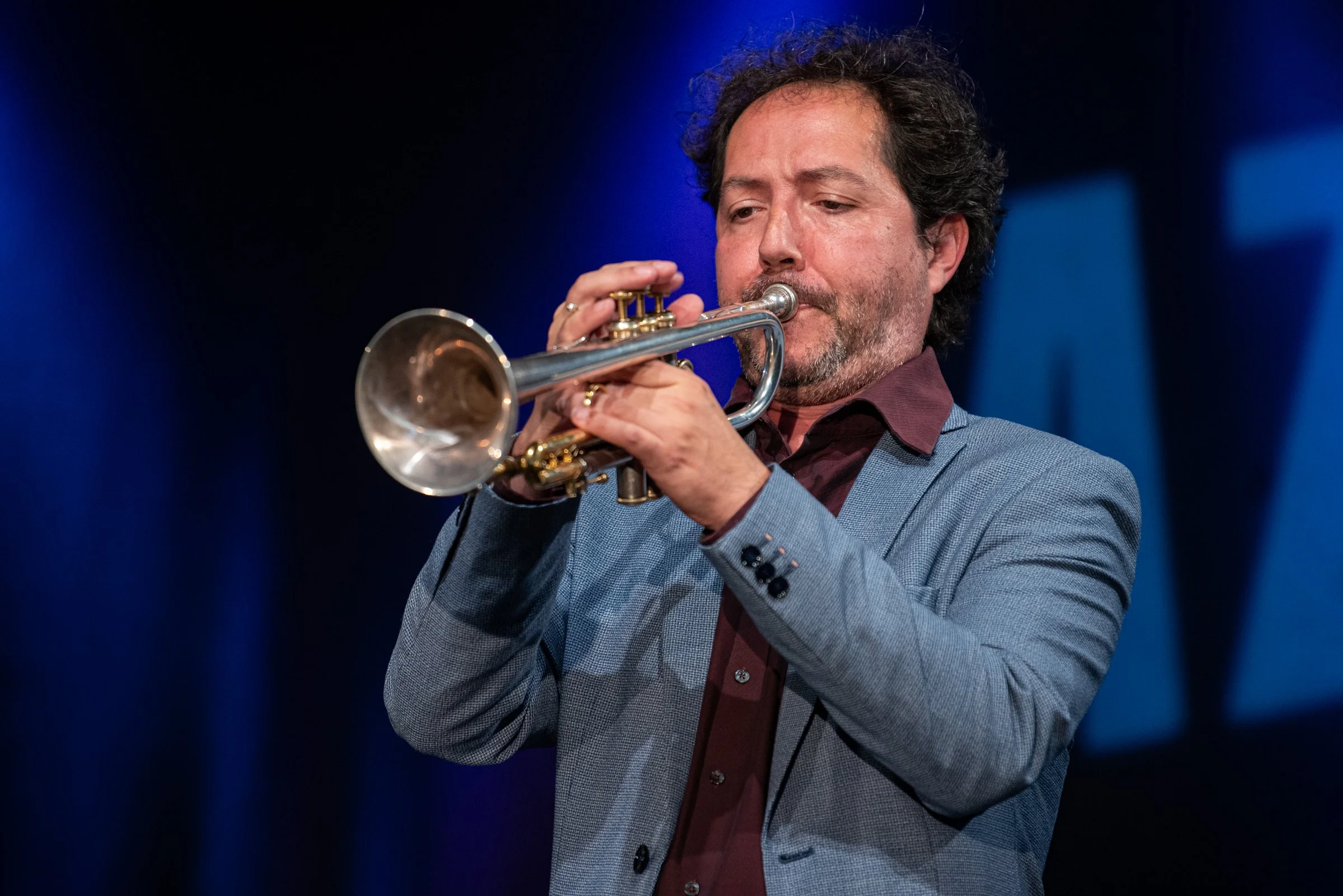 Ein Mann spielt auf einer Trompete, trägt einen grauen Blazer und hat dunkle, lockige Haare, steht vor einem dunklen Hintergrund.