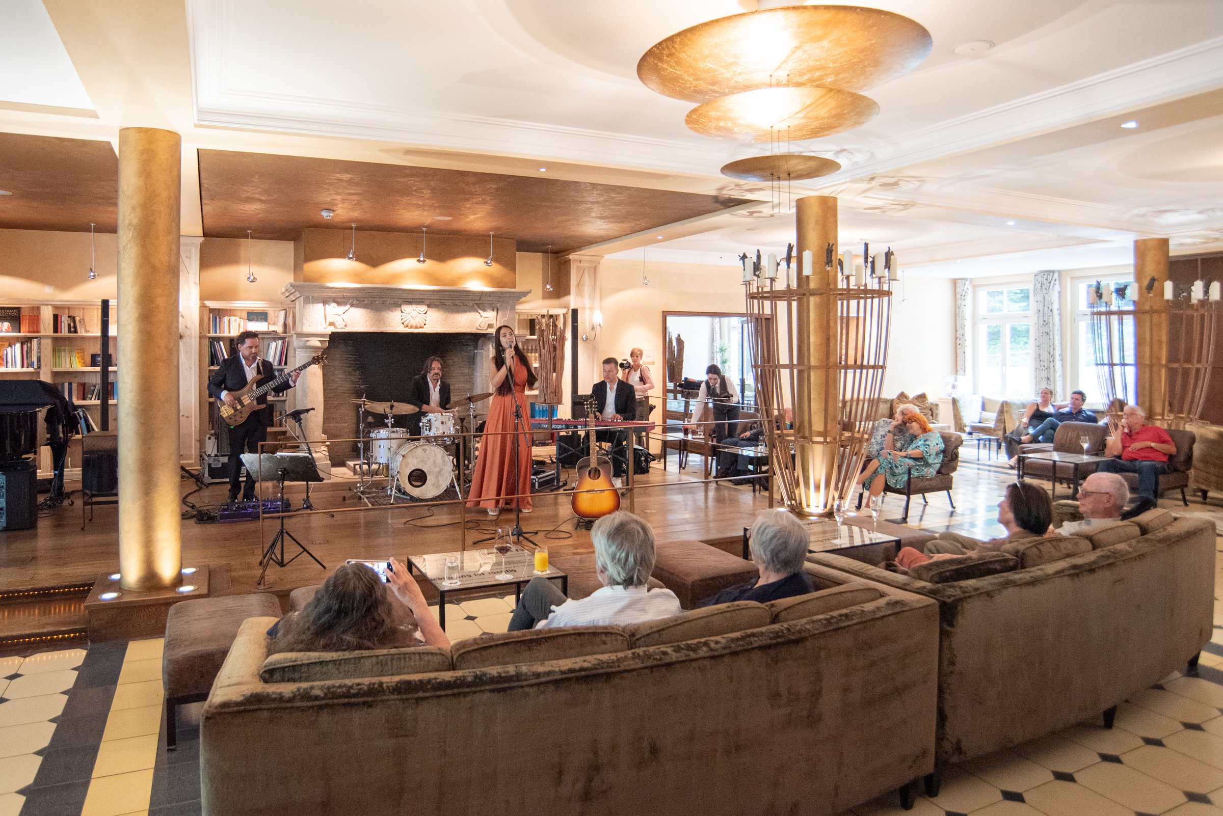 Eine Band spielt in einem eleganten Hotelraum, während das Publikum auf gemütlichen Sofas und Stühlen sitzt und zuhört.