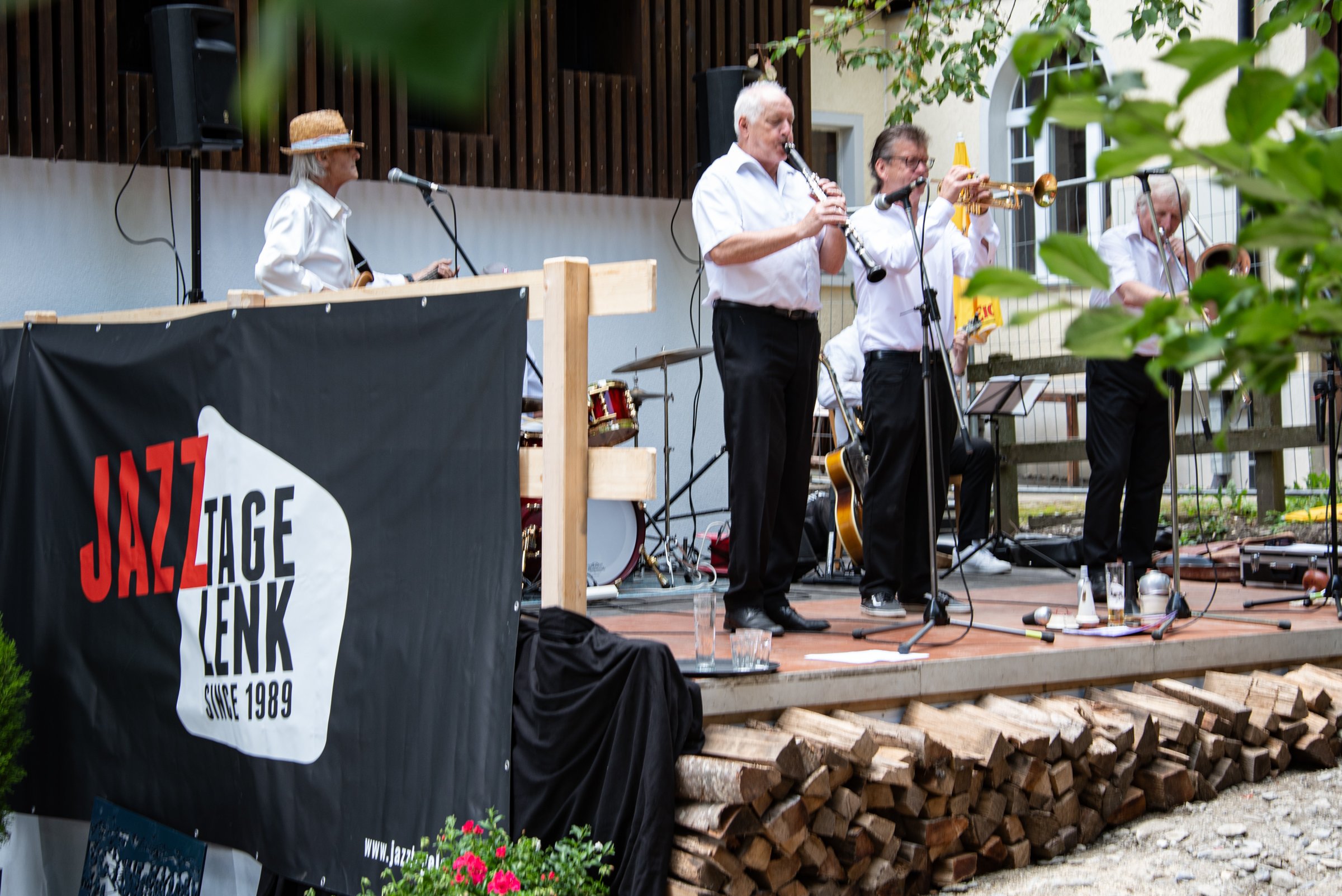 Eine Band spielt auf einer kleinen Bühne im Freien. Die Musiker tragen weiße Hemden und schwarze Hosen. Es sind mehrere Musiker, darunter ein Klarinettist, ein Trompeter und andere Instrumentalisten. Im Vordergrund ist ein schwarzes Banner mit dem Te