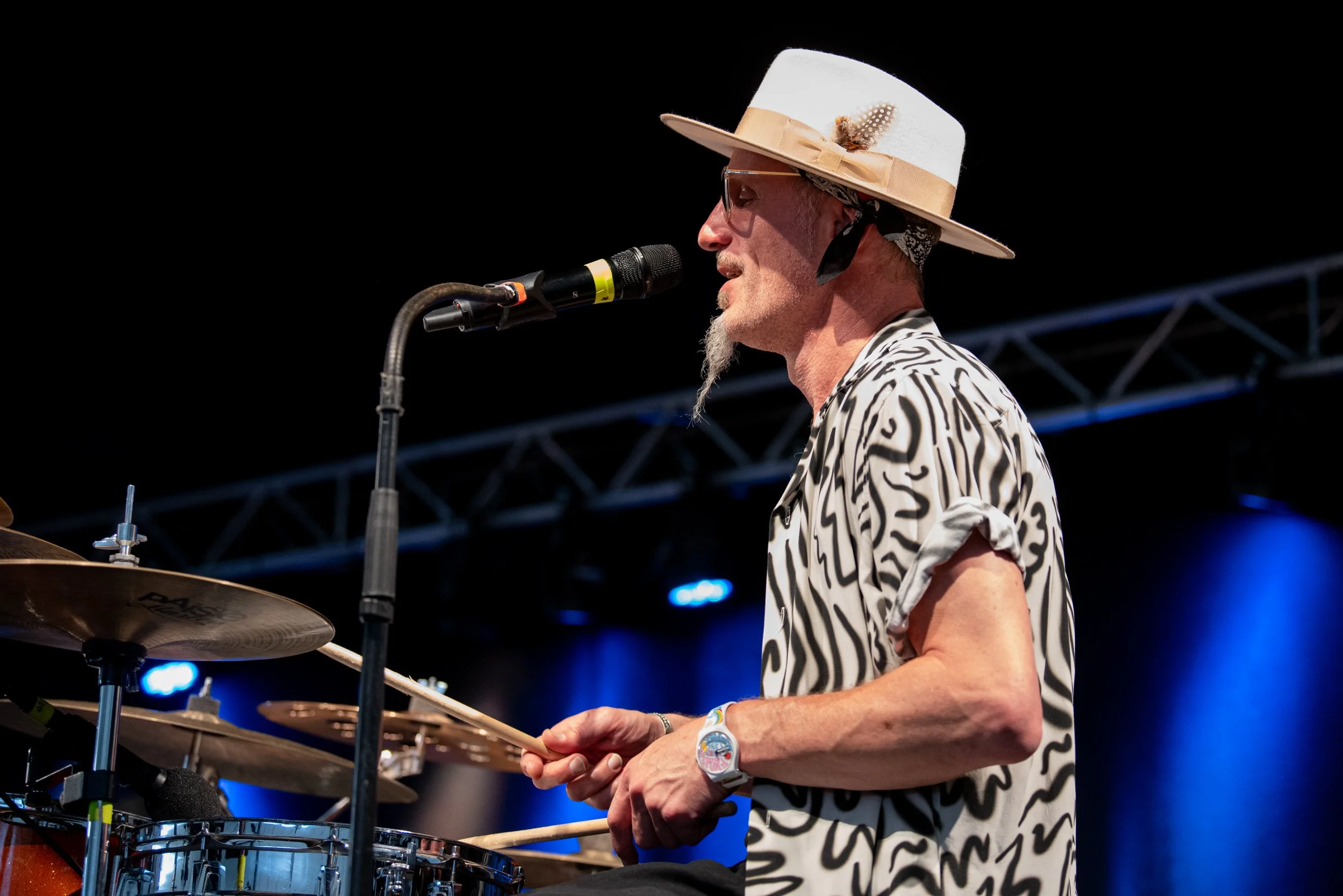 Ein älterer Mann spielt Schlagzeug auf einer Bühne, trägt einen Sonnenhut mit Schleife und Feder, eine gestreifte Bluse, eine Uhr, und befindet sich vor einem Mikrofon vor dunklem Hintergrund mit blauen Lichtern.