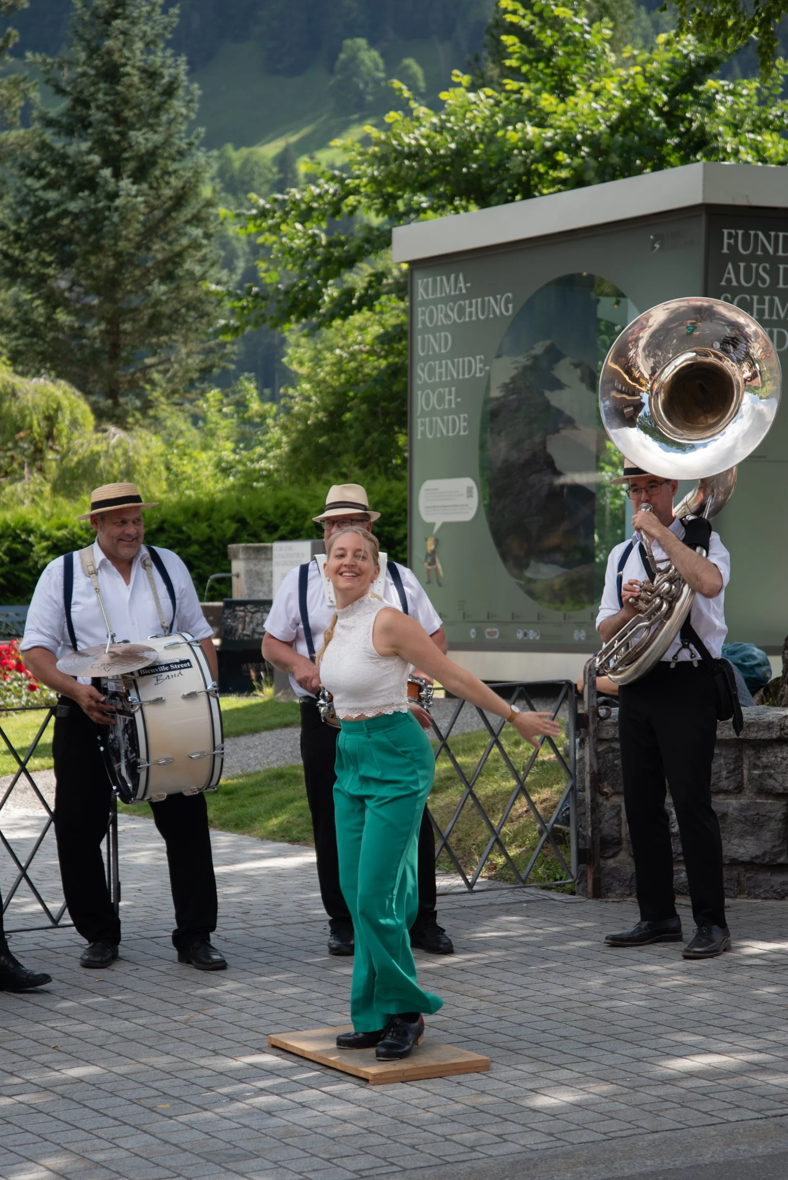 Musiker spielen auf einer Straße mit einer Frau, die auf einem Holzblock tanzt, im Hintergrund ein Informationsschild und grüne Bäume.