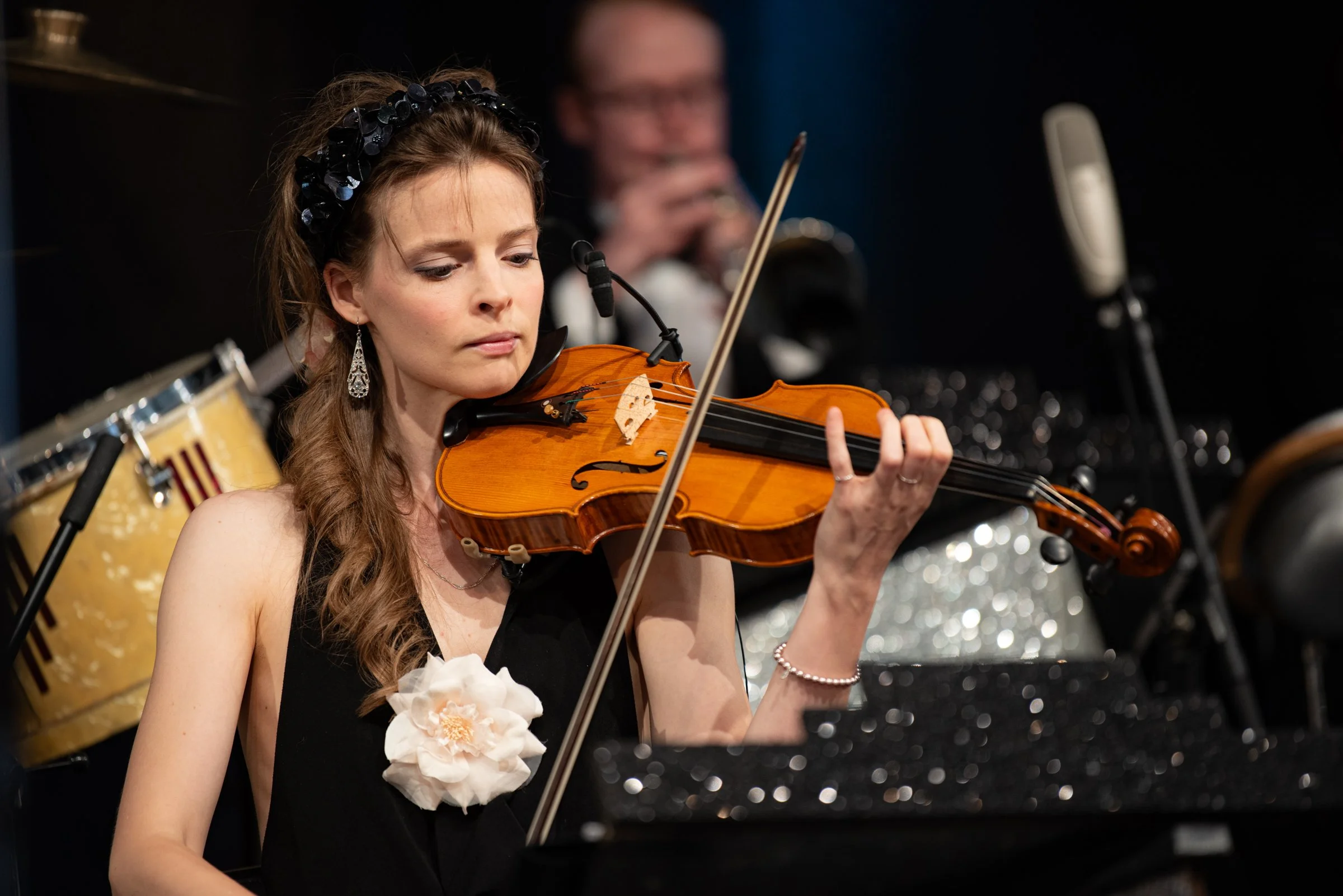 Eine Frau spielt Geige bei einem Orchesterauftritt, im Hintergrund sind weitere Musiker und ein Schlagzeug zu sehen.