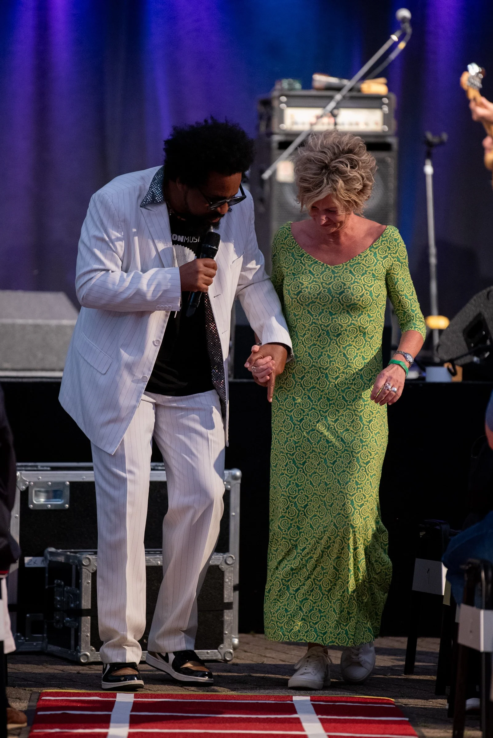 Zwei Menschen auf einer Bühne, die sich die Hand halten und sich verabschieden, während im Hintergrund musikalische Geräte stehen.
