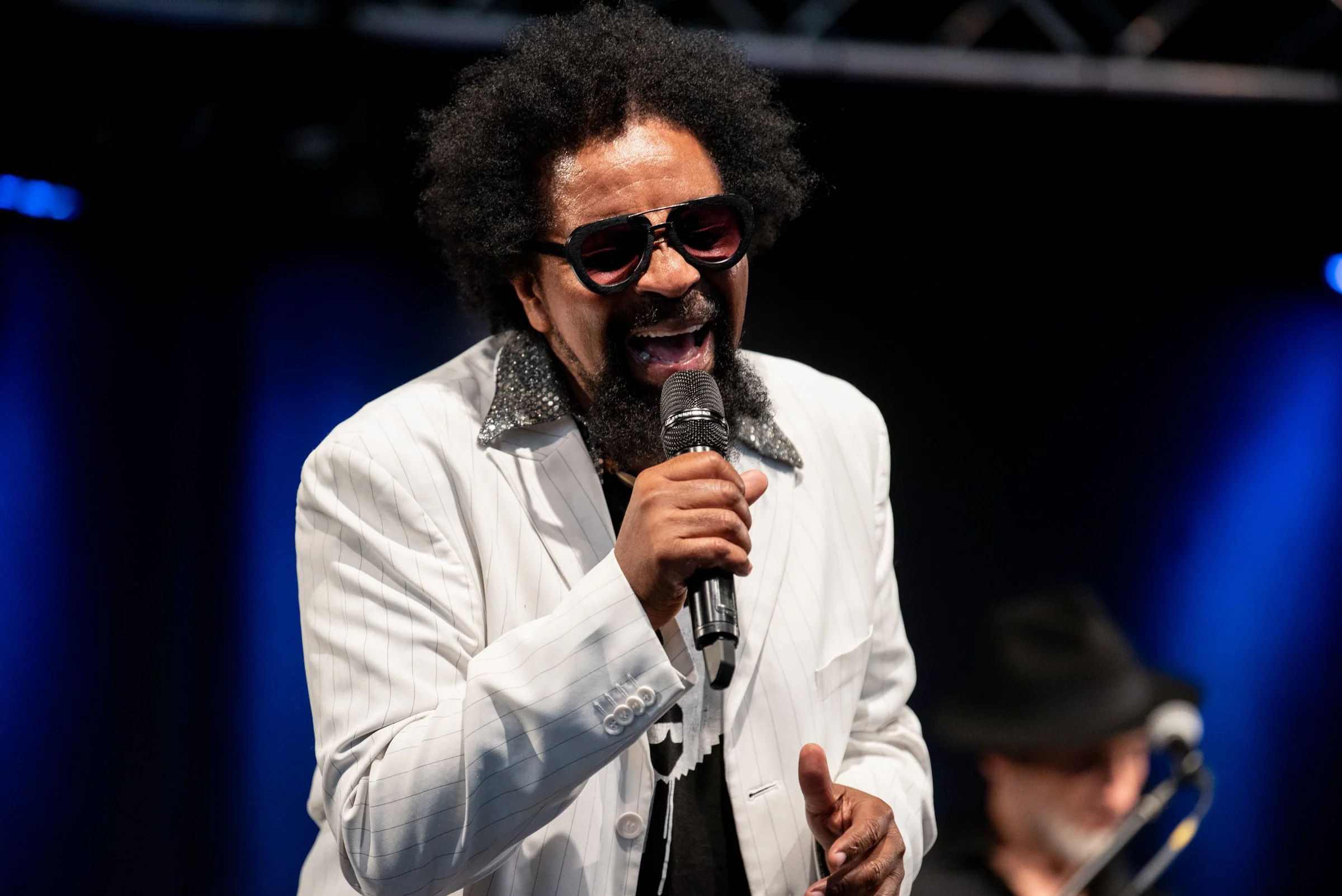 Ein Sänger mit dunkler Haut, afroamerikanischer Frisur, Sonnenbrille und weißen Anzug mit glänzender Kragenstickerei singt in einem dunklen Raum mit blauer Beleuchtung, während im Hintergrund eine Person mit Hut Gitarre spielt.