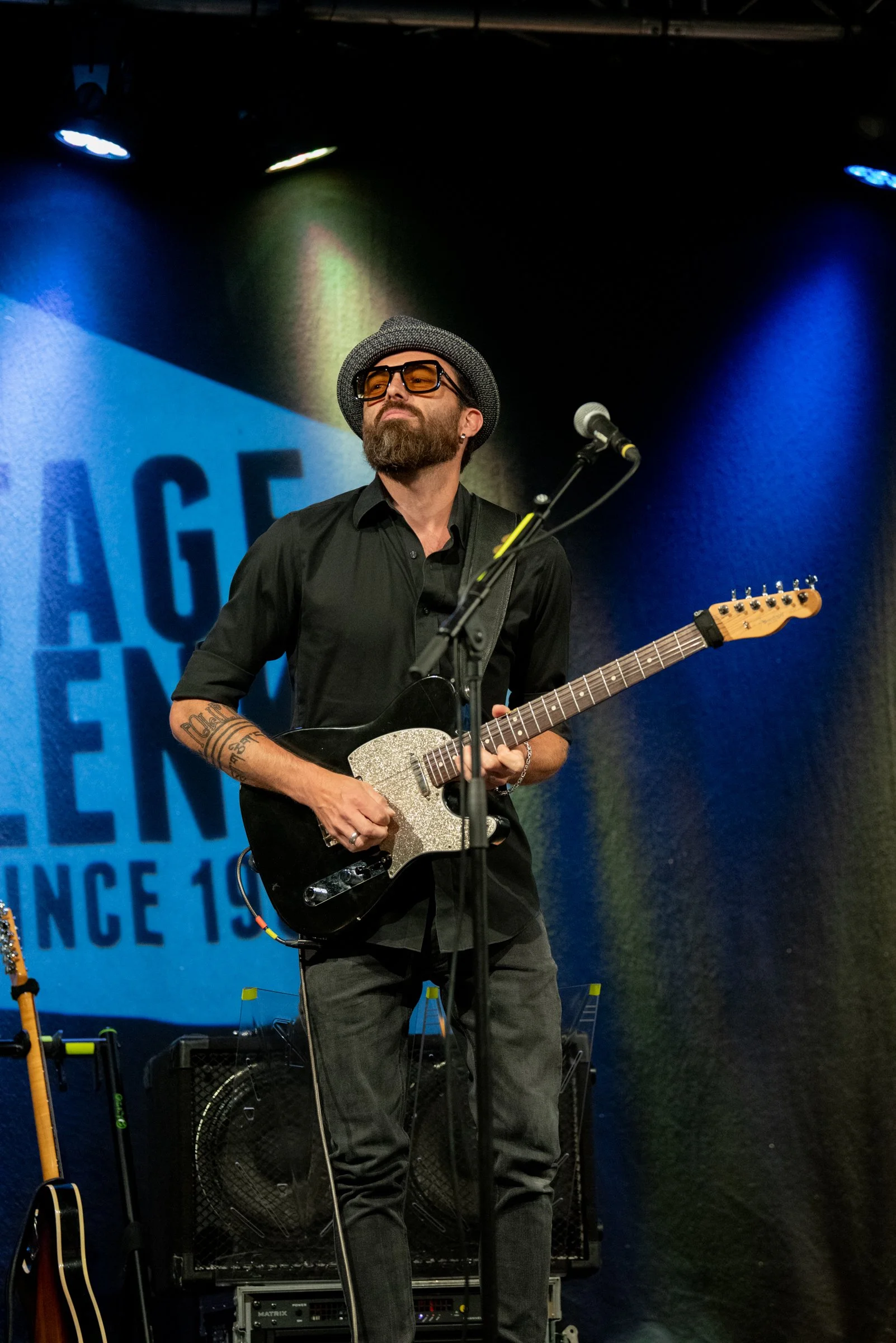 Mann mit schwarzen Hemd, Brille, Hut und tätowierten Armen spielt Gitarre auf Bühne bei einem Konzert.