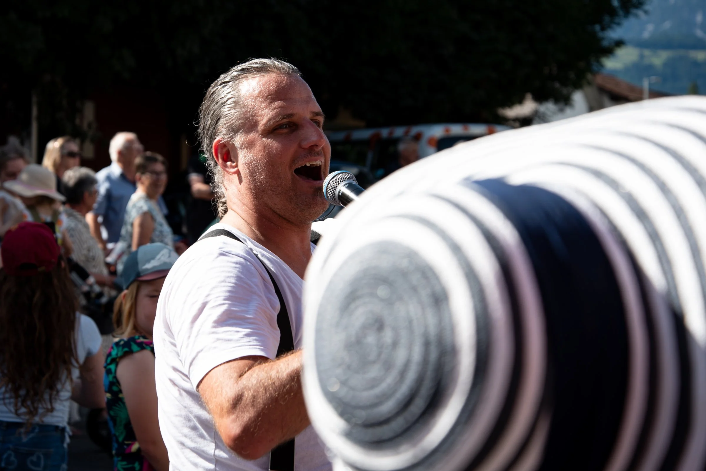Mann spricht in einem Mikrofon bei einem öffentlichen Event im Freien, im Hintergrund Menschen, einige Kinder, die zuschauen.