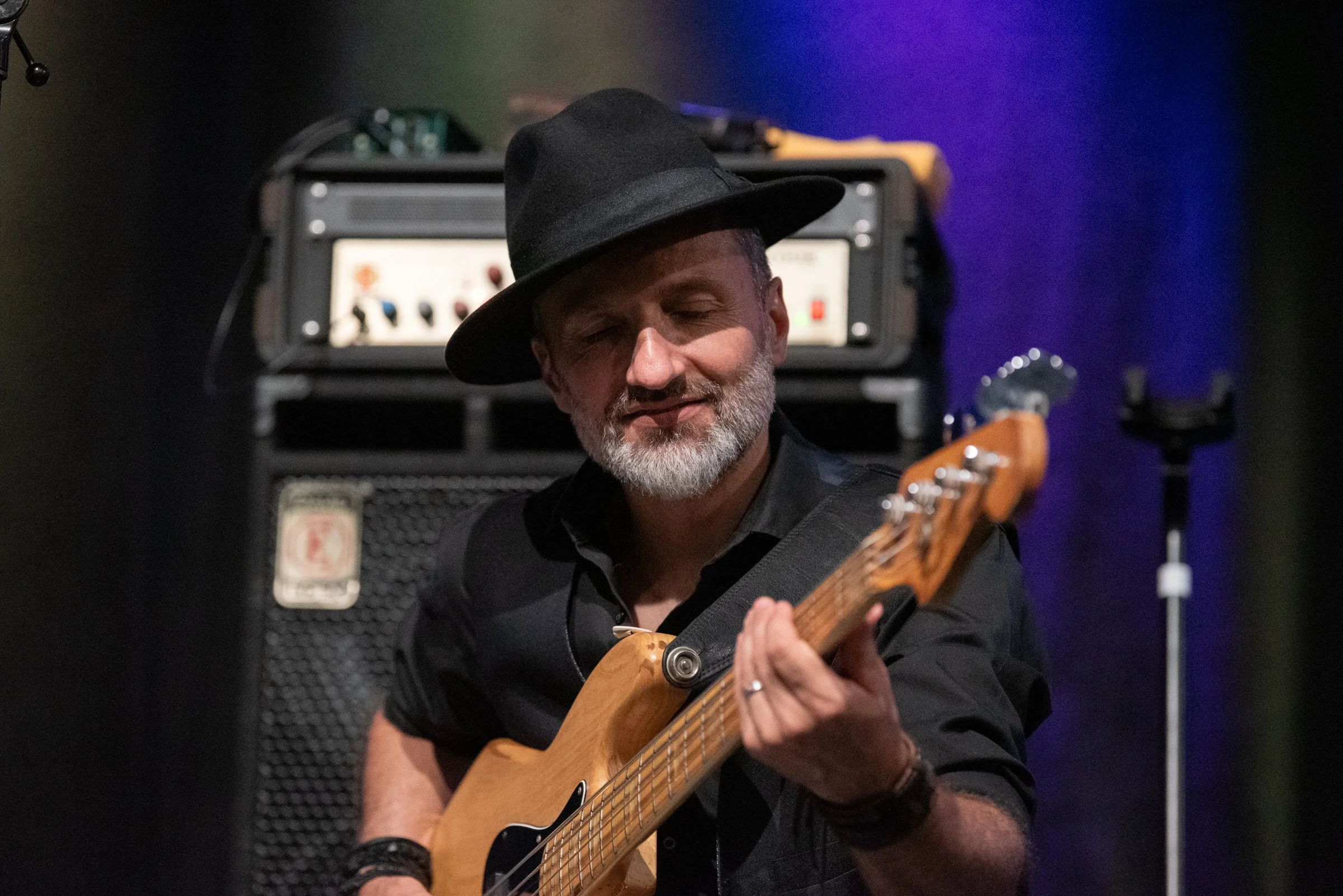 Mann spielt Gitarre auf Bühne, trägt schwarzen Hut und dunkle Kleidung, im Hintergrund Verstärker und blaues Licht.