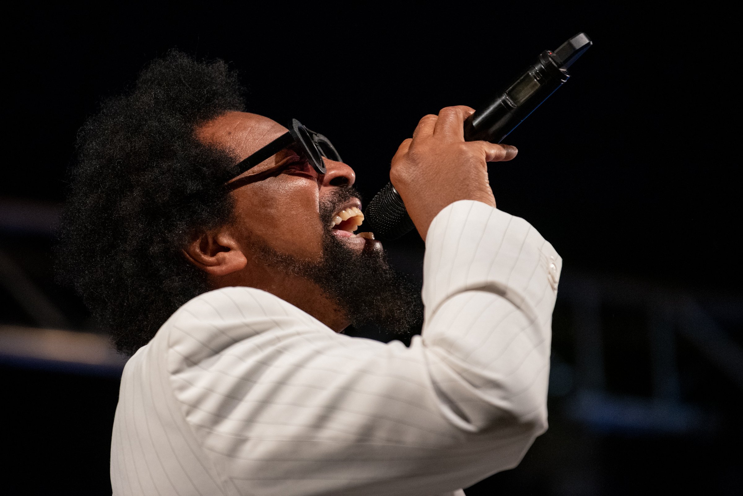 Ein Mann mit dunkler Haut, schwarzen lockigen Haaren, Sonnenbrille und weißem Anzug singt in einem Karaoke-Mikrofon bei dunklem Hintergrund.