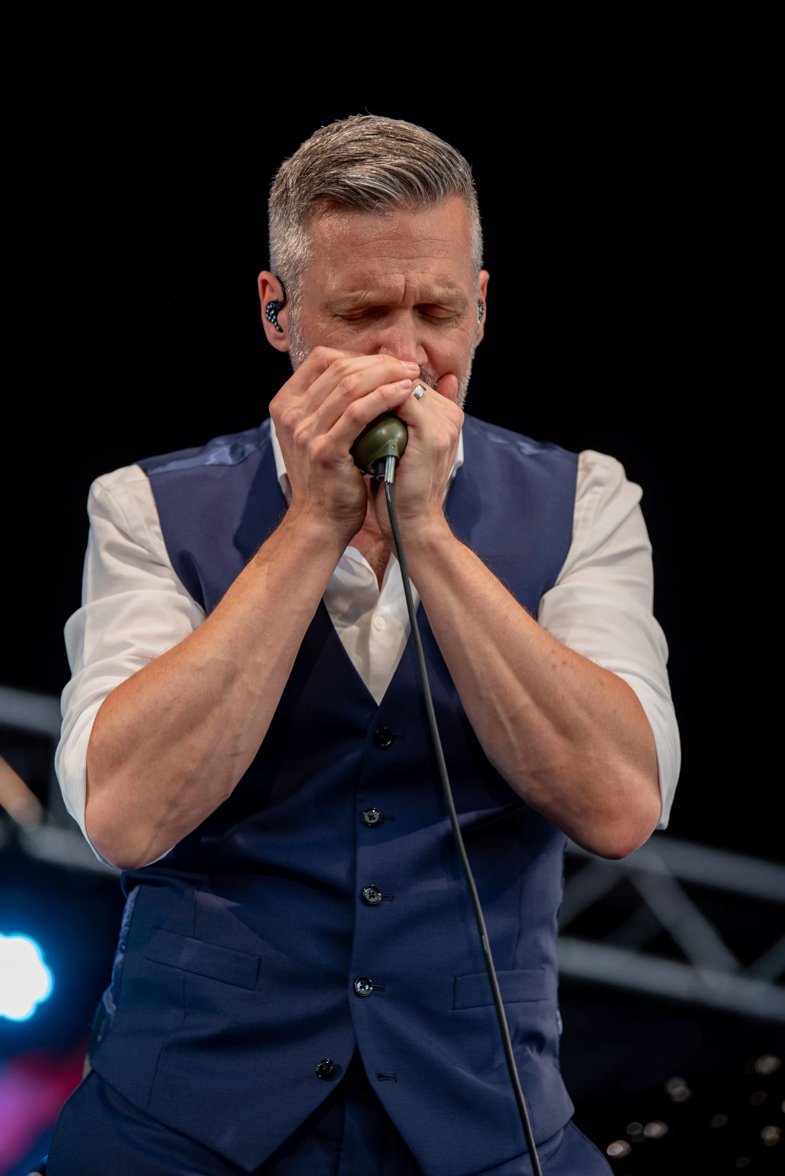 Mann spielt Harmonika bei einem Auftritt, trägt einen dunklen Anzug mit weißen Hemd, Blick nach unten, Hintergrund dunkel.