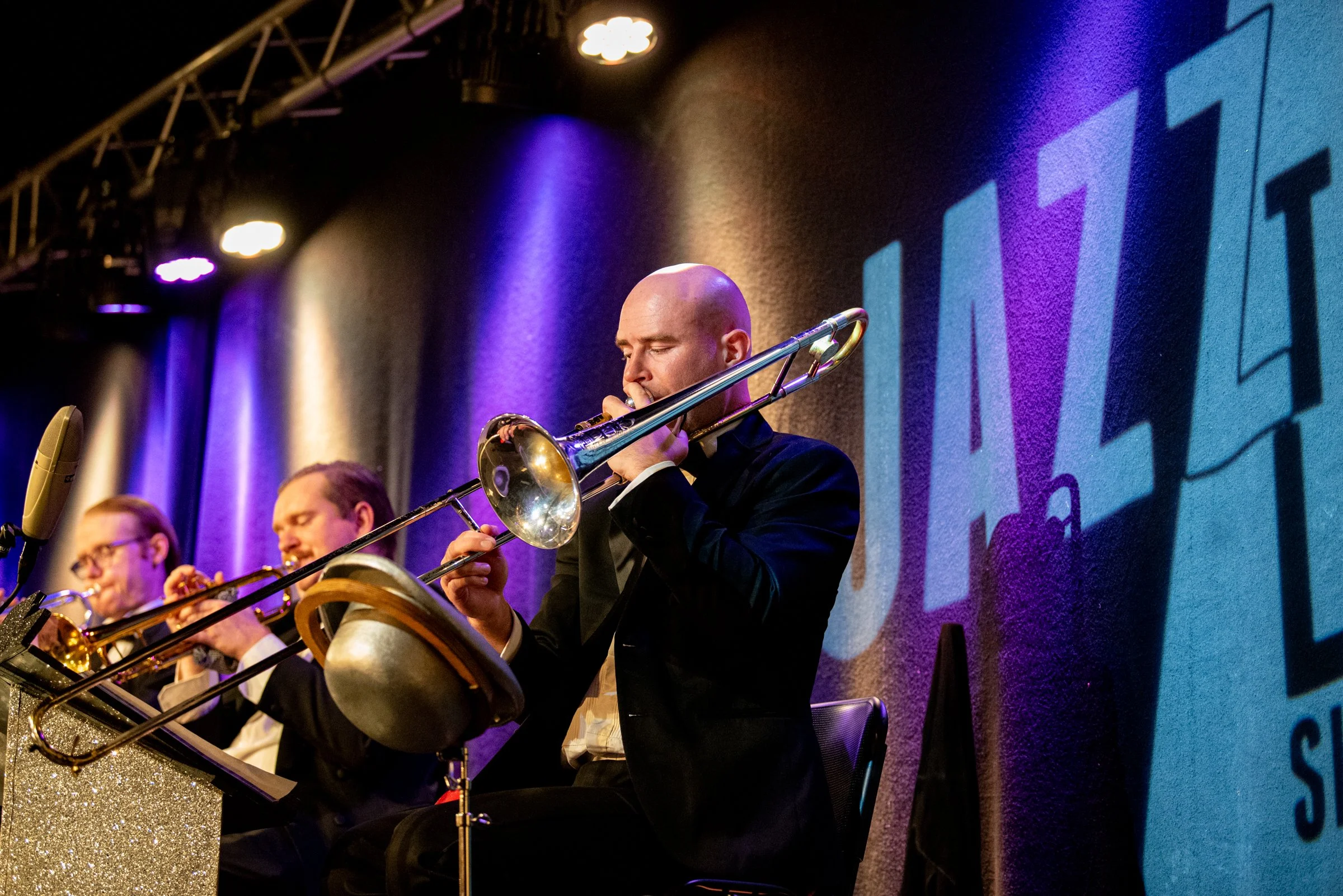 Musiker spielt Trompete bei einem Konzert, im Hintergrund ein großes Logo mit der Aufschrift 'JAZZ' an einer Bühne mit buntem Licht.