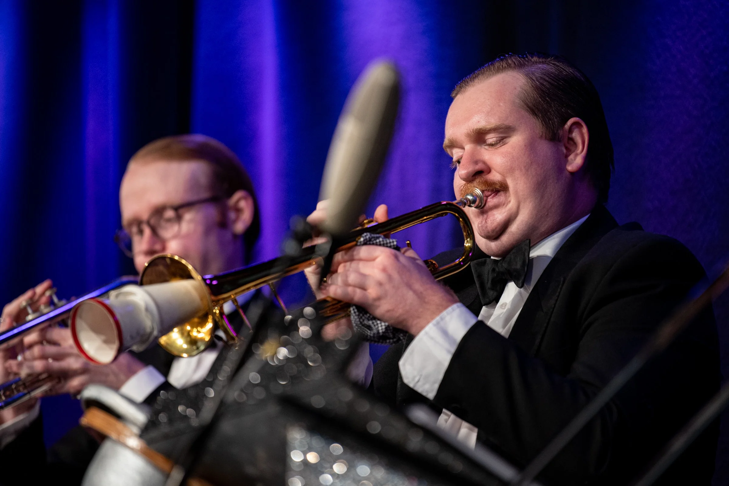 Zwei Männer spielen Trompete in formeller Kleidung bei einem Konzert, blauer Bühnenhintergrund.