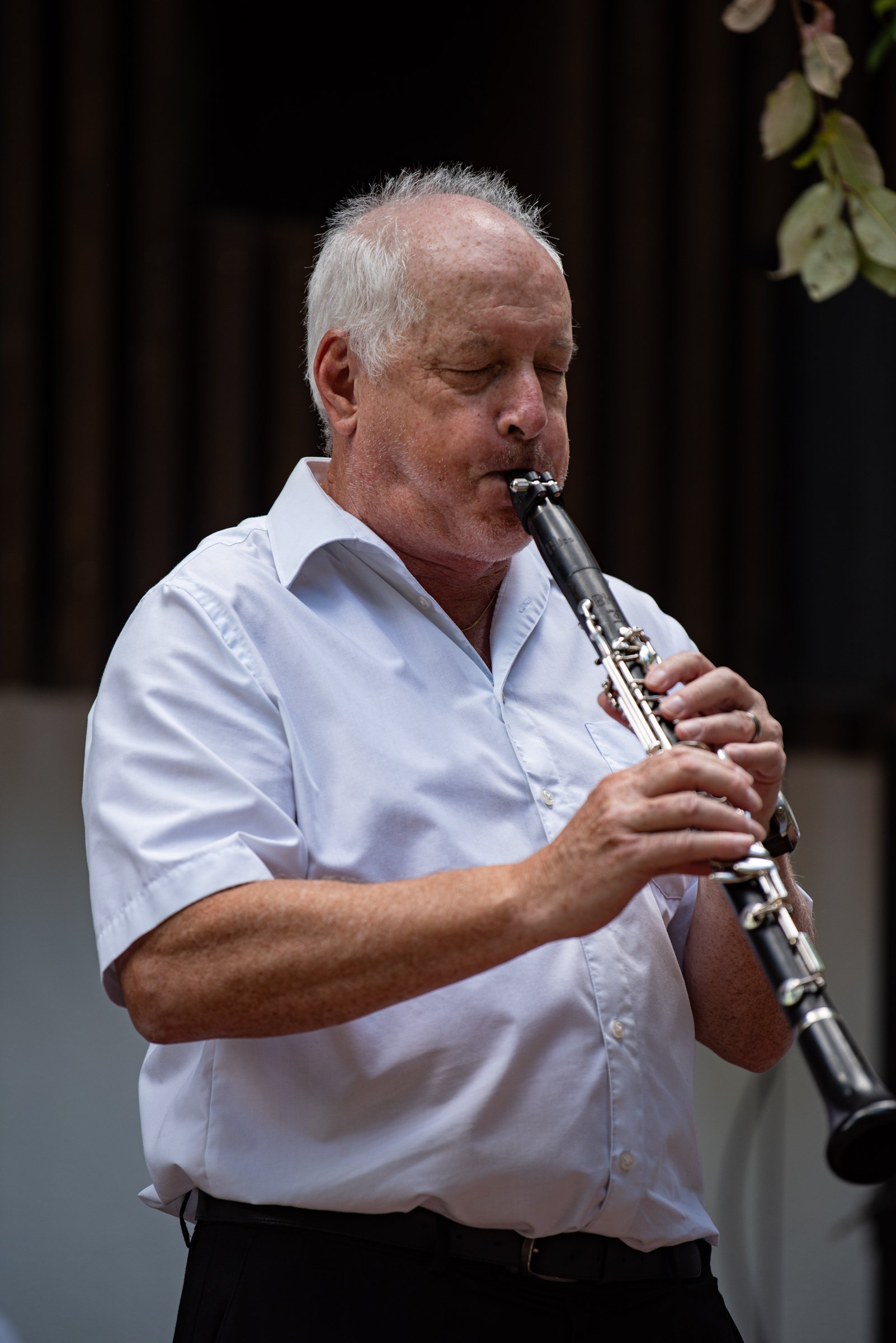 Ein älterer Mann spielt auf einem schwarzen Klarinette. Er trägt ein weißes Hemd und hat kurze graue Haare.