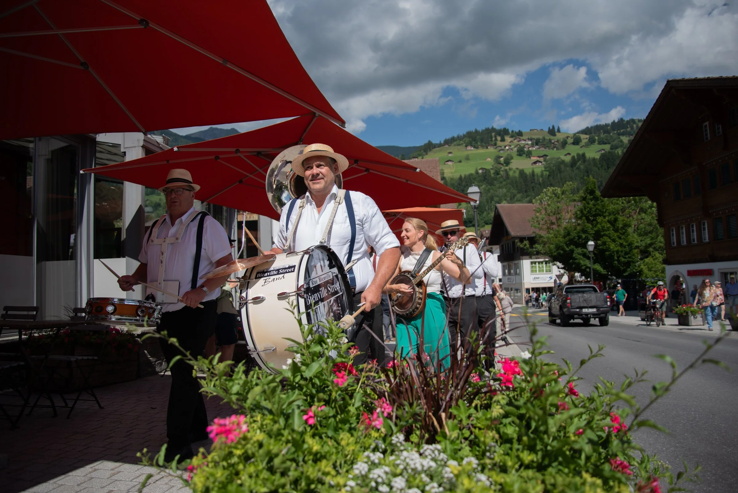 Eine Gruppe von Musikern in traditionellen Trachten spielt auf der Straße in einer malerischen Stadt, umgeben von Bergen und bunten Blumen.