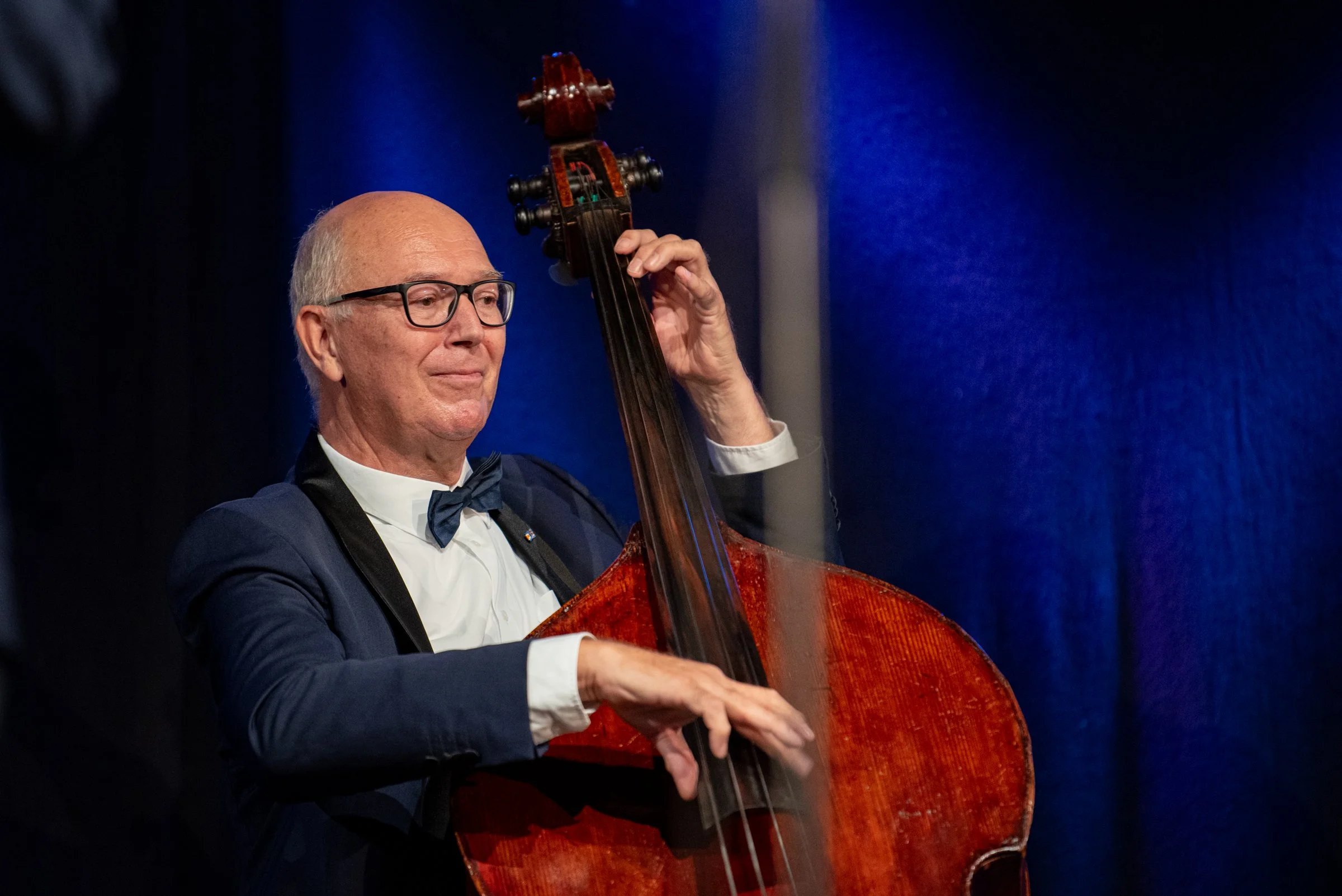 Ein älterer Mann trägt einen schwarzen Anzug mit Fliege und spielt auf einem Kontrabass auf einer Bühne mit blauer Hintergrundbeleuchtung.