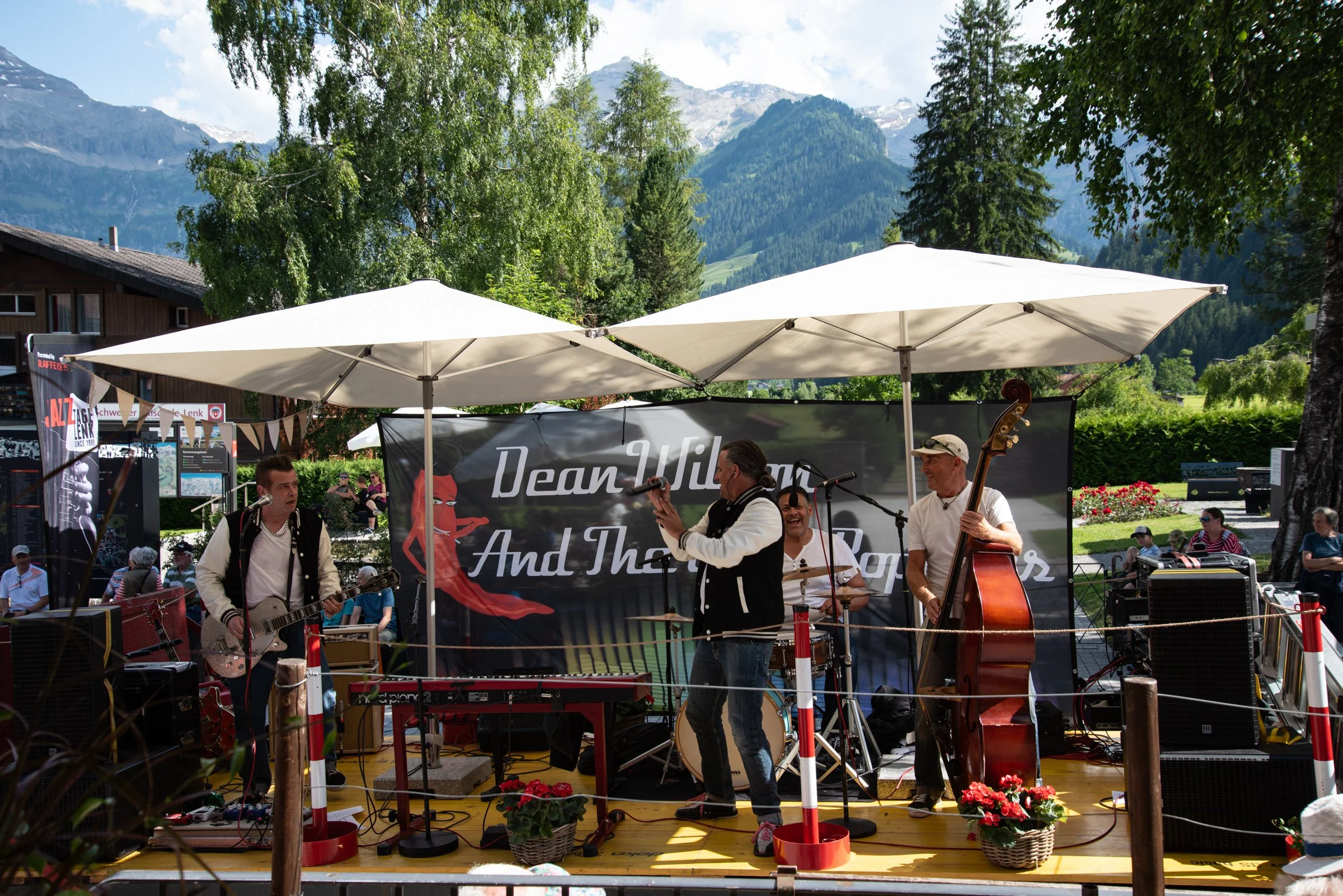 Eine Band spielt auf einer Bühne im Freien, umgeben von Bergen und grüner Natur, bei sonnigem Wetter. Die Musiker bestehen aus einem Gitarristen, einem Sänger, einem Schlagzeuger und einem Bassisten. Es gibt Menschen im Publikum, die die Musik genieß