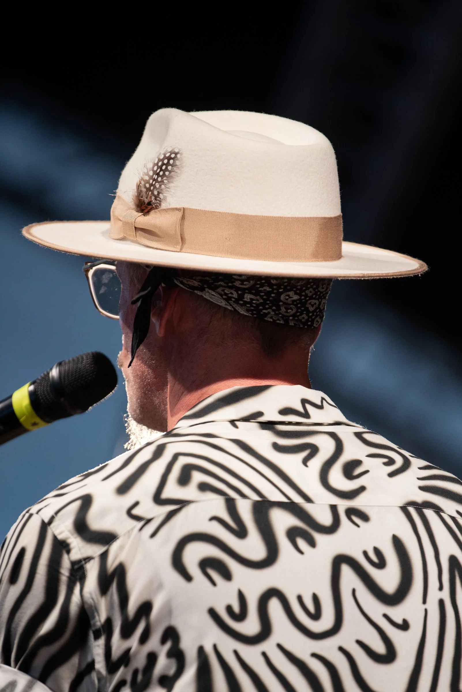 Mann mit weißem Hut mit beige Band, schwarzem Halstuch, Brille und muskulösem Rücken mit schwarzem und weißem Muster, spricht in ein Mikrofon.