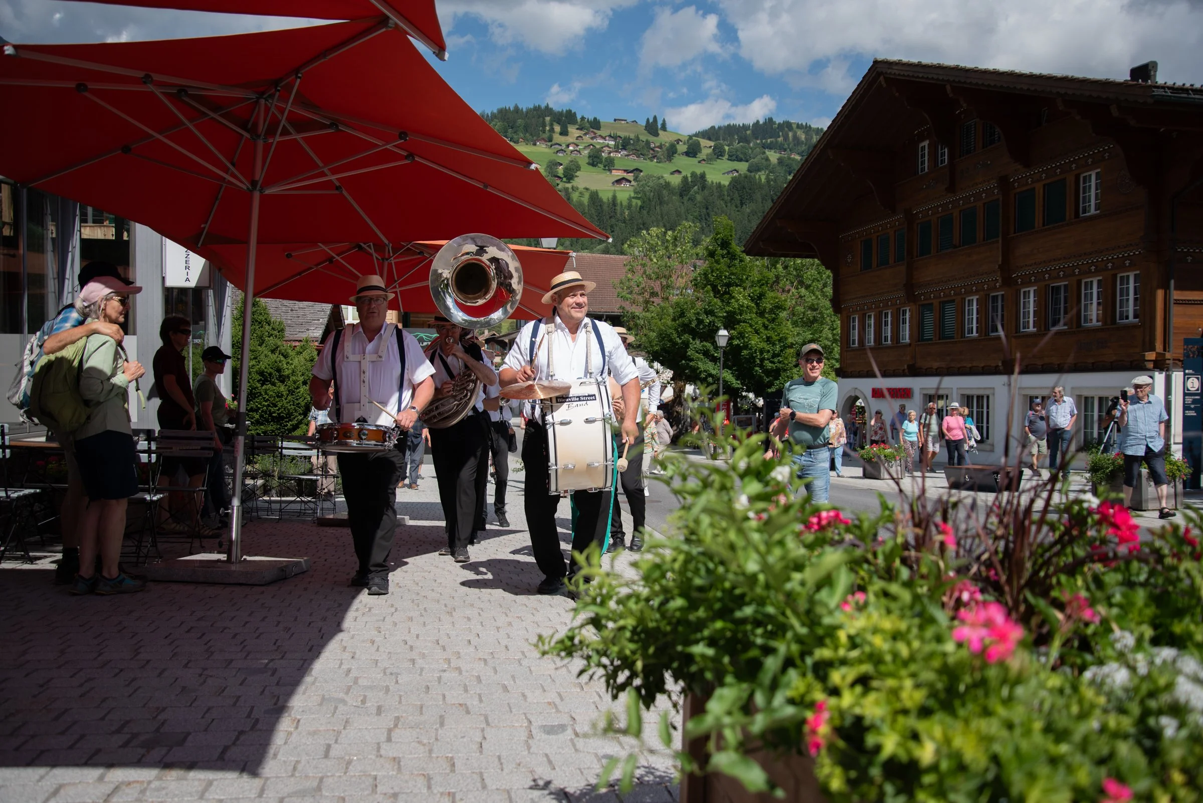 Eine Straßenband spielt im Freien vor einem roten Sonnenschirm, umgeben von Passanten in einer malerischen Stadt mit grünen Hügeln im Hintergrund. Es ist ein sonniger Tag.
