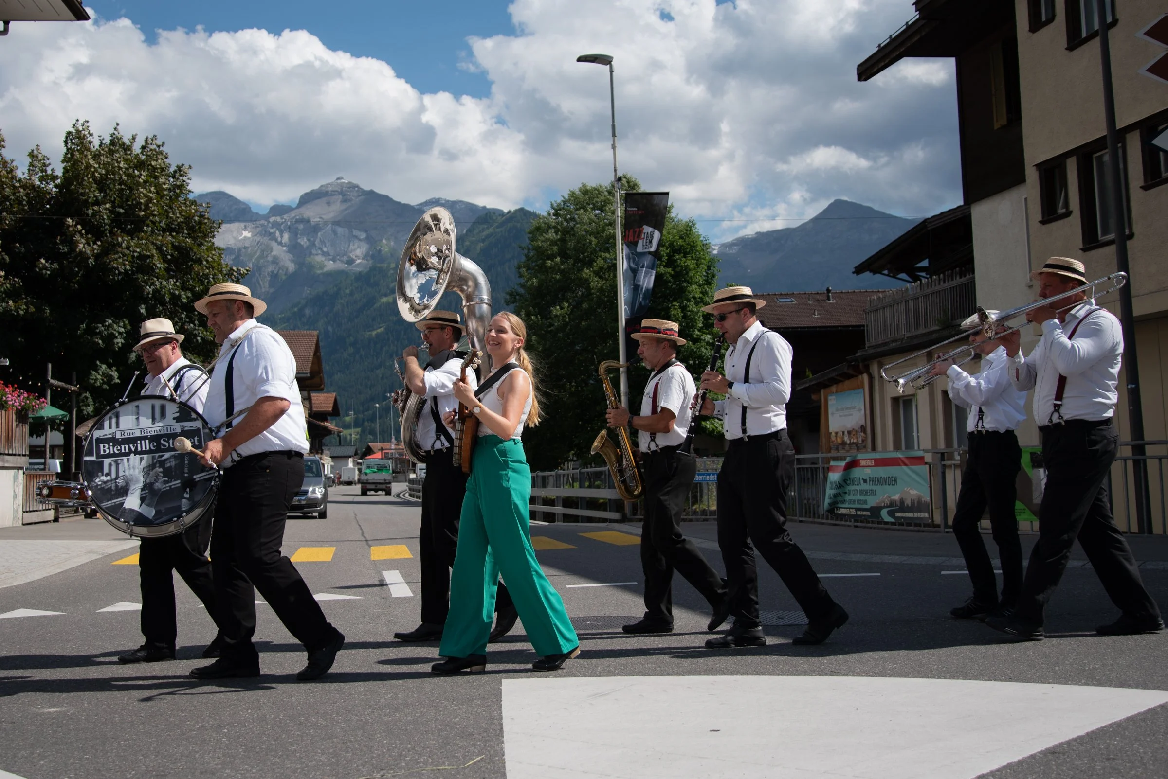 Eine Band bestehend aus mehreren Mitgliedern spielt auf einer Straße im Freien. Sie trägt weiße Hemden, schwarze Hosen, und Strohhüte. Einige spielen Blechblasinstrumente wie Trompete und Tuba, während eine eine Gitarre spielt und eine andere eine gr
