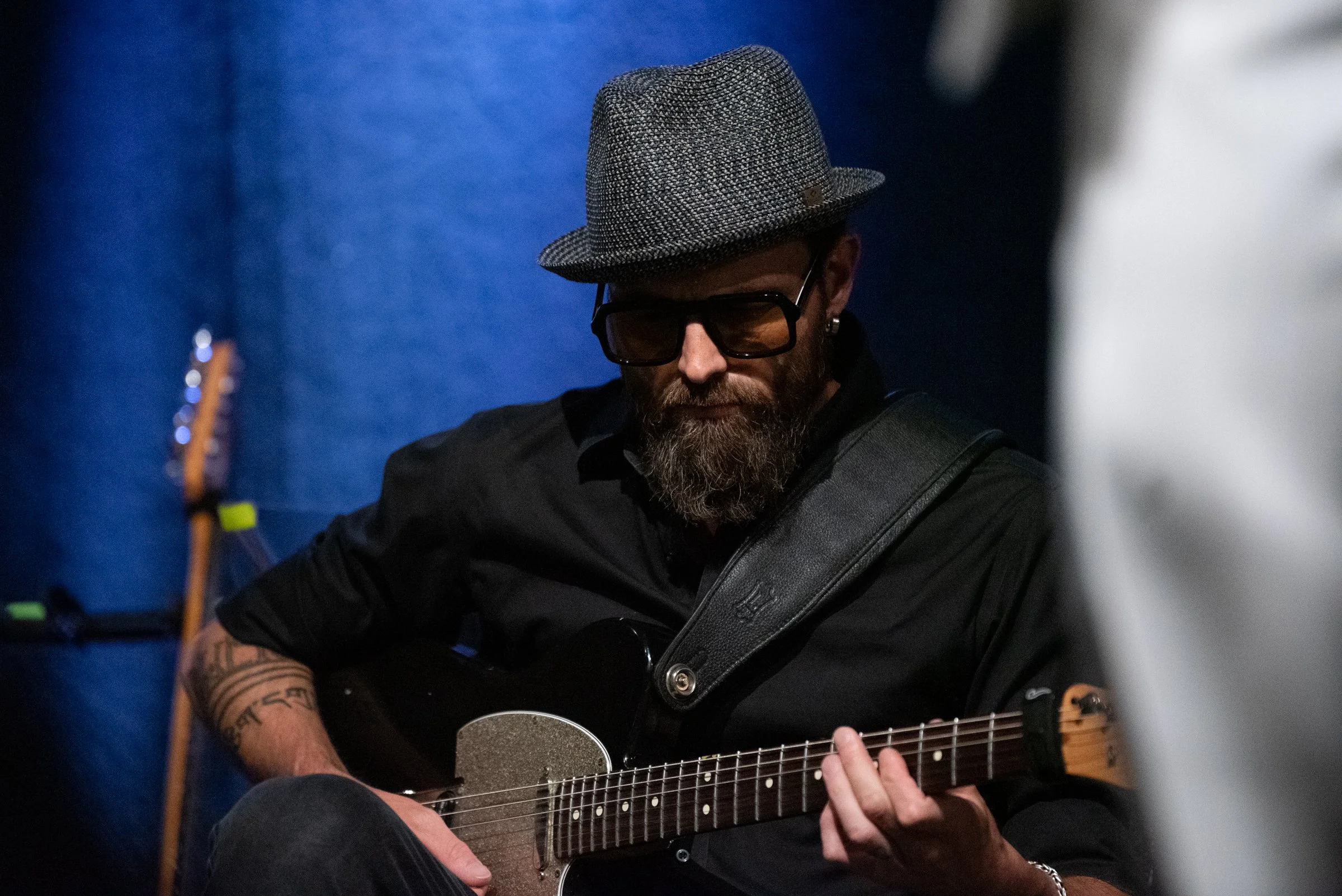 Mann mit Hut und Brille spielt Gitarre in dunklem Raum.