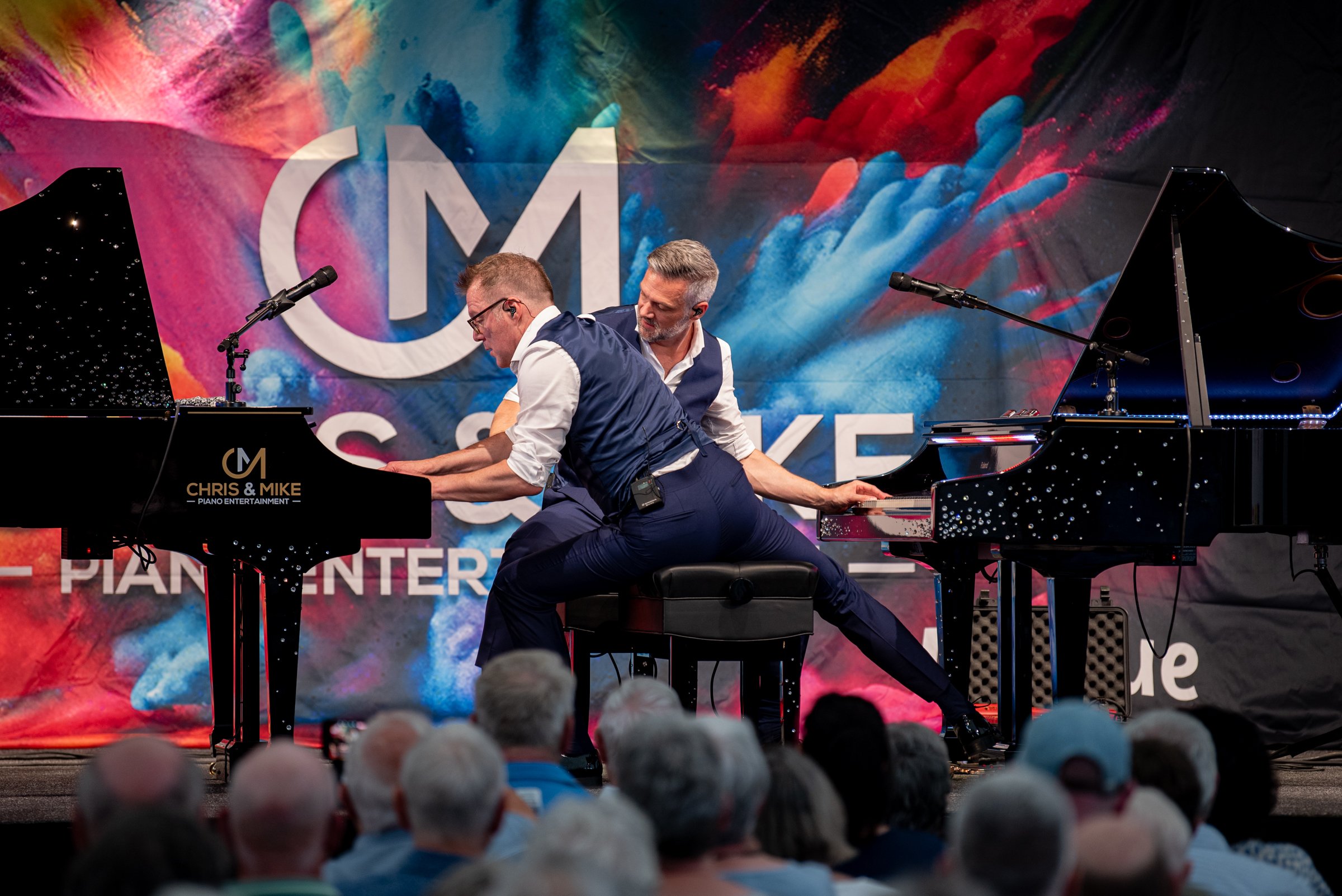 Zwei Musiker spielen auf beiden Klavieren während eines öffentlichen Konzerts. Der Hintergrund zeigt ein farbenfrohes, abstraktes Design mit den Worten "Chris & Mike Piano Entertainment".