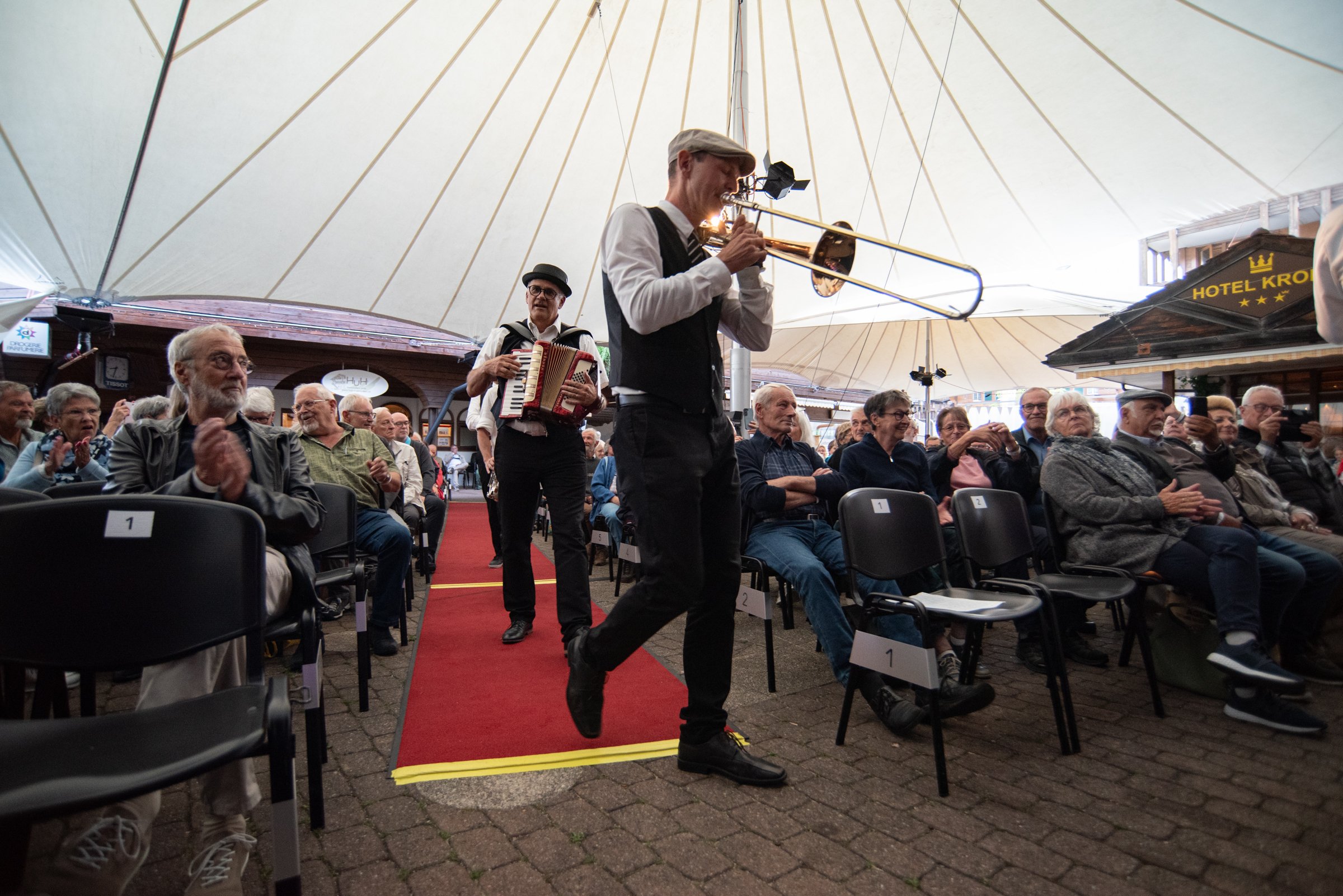 Musiker spielen im Zelt vor einem Publikum von alten Menschen, das sitzend zuschaut. Der Musiker im Vordergrund spielt Trompete, während ein weiterer Musiker mit Akkordeon im Hintergrund steht. Das Zelt ist groß und hat eine große, helle Decke, und e