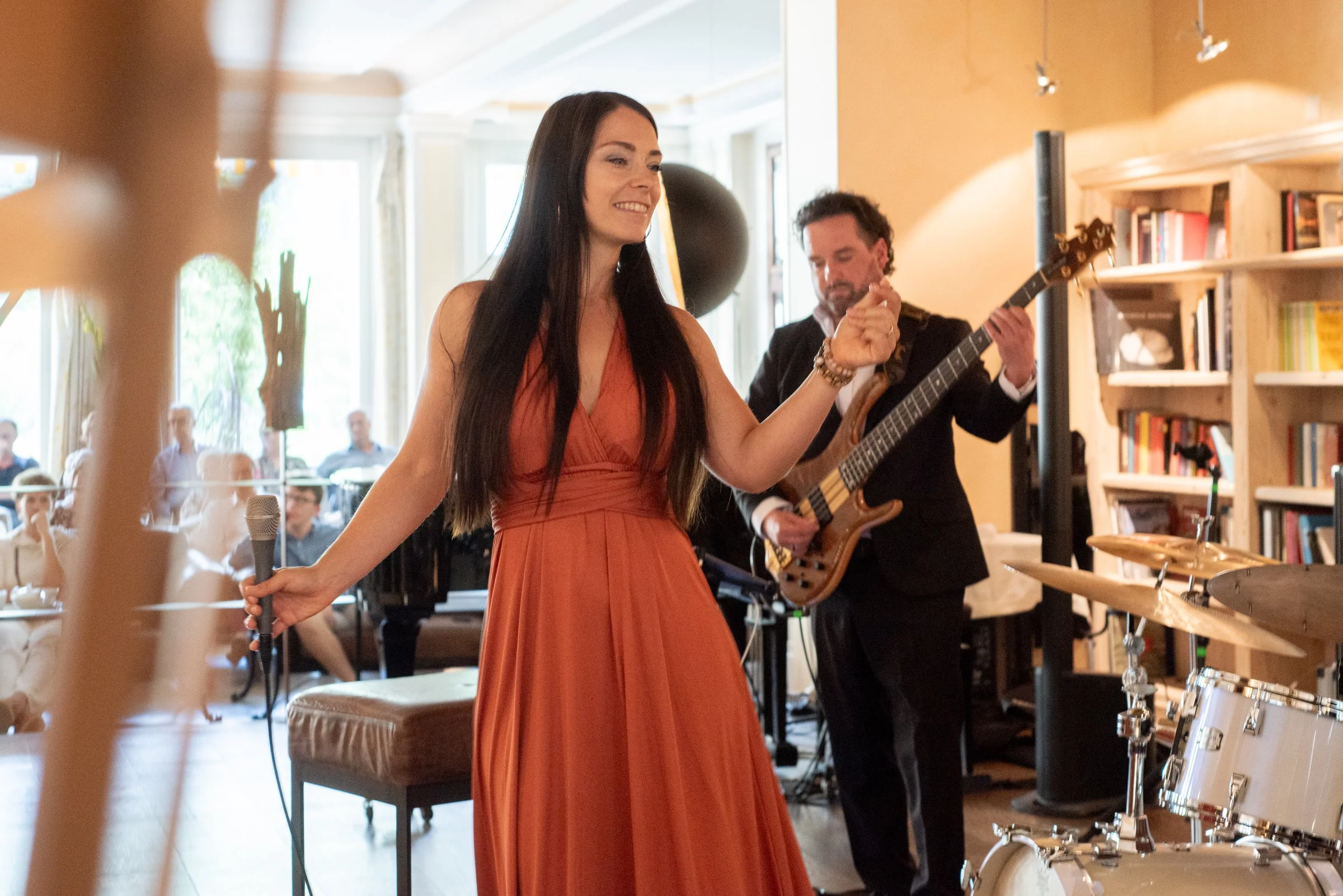 Eine Frau in einem roten Kleid hält ein Mikrofon in der Hand, während ein Mann im schwarzen Anzug Gitarre spielt. Im Hintergrund sitzen Gäste in einem gemütlichen Raum.