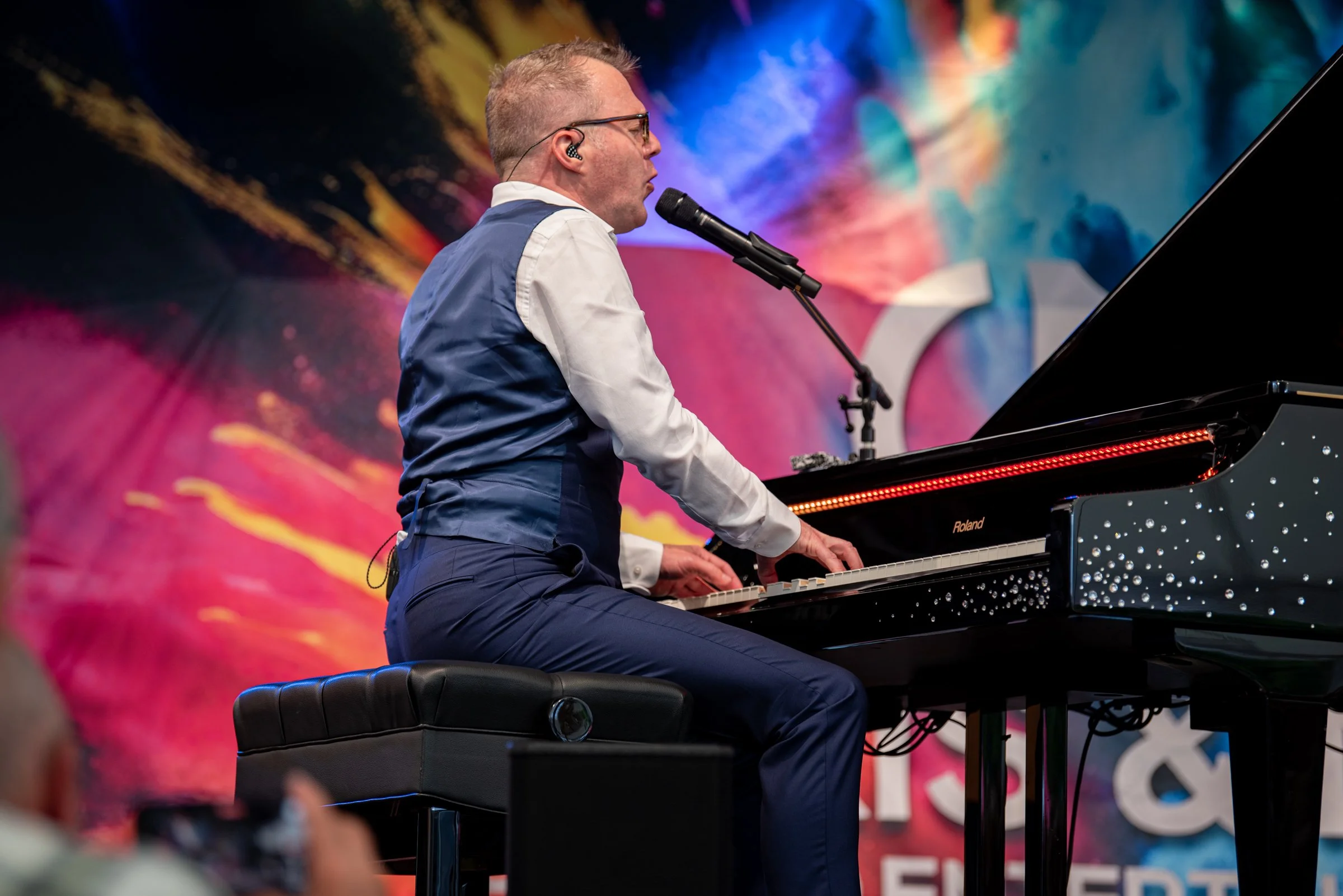 Ein Mann spielt Klavier auf einer Bühne mit buntem Hintergrund, trägt Anzug und trägt ein Headset.