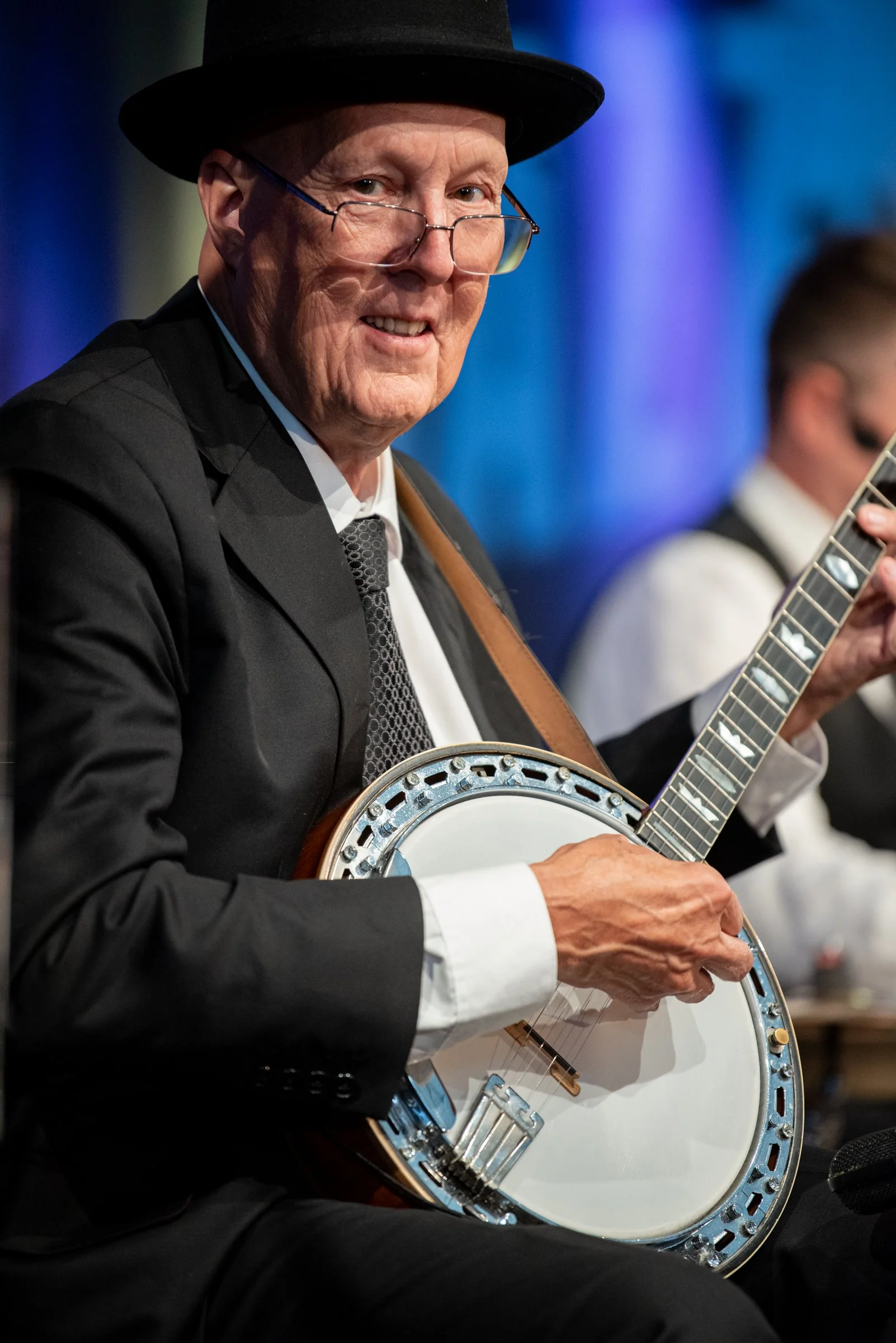 Ein älterer Mann trägt einen schwarzen Anzug, ein weißes Hemd, eine dunkle Krawatte und eine schwarze Hut und spielt Gitarre bei einem Musikauftritt.