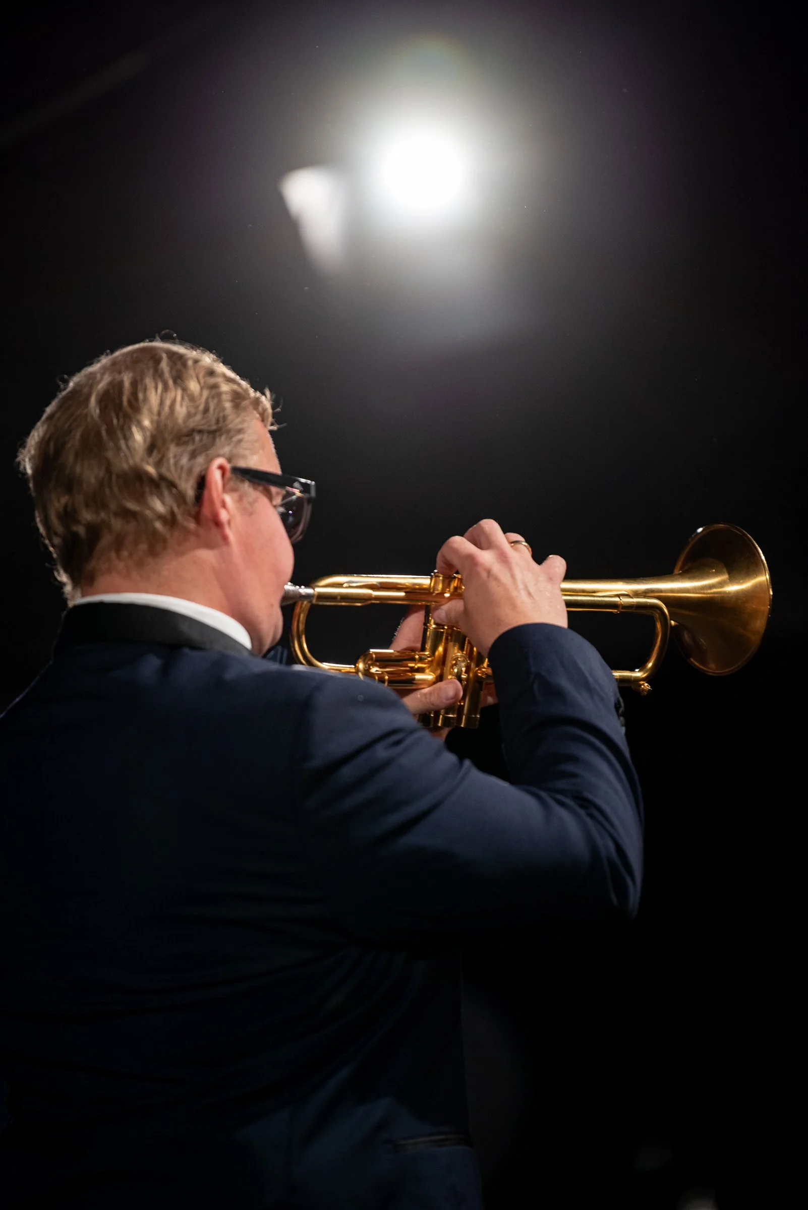Ein Mann in einem dunklen Anzug spielt auf einem goldenen Trompete gegen einen dunklen Hintergrund, beleuchtet von einer starken Lichtquelle im oberen Bereich.