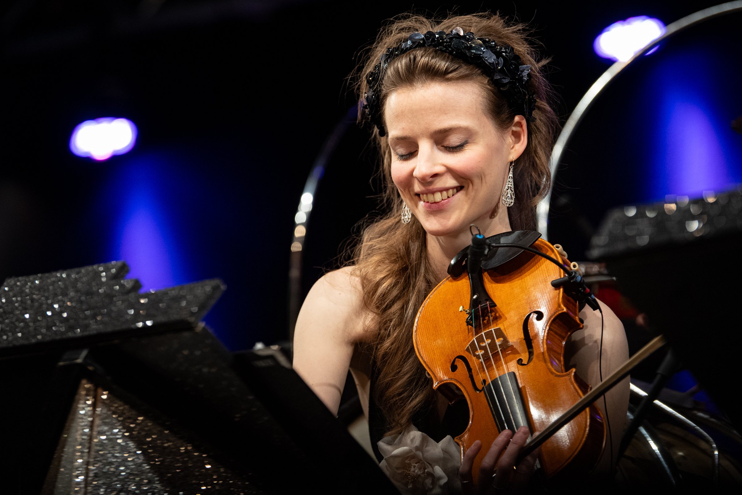Junge Frau spielt Geige auf einer Bühne, trägt Ohrringe und ein schwarzes Kleid, hat braune Haare und ein schwarzes Stirnband, im Hintergrund blaue Lichter.