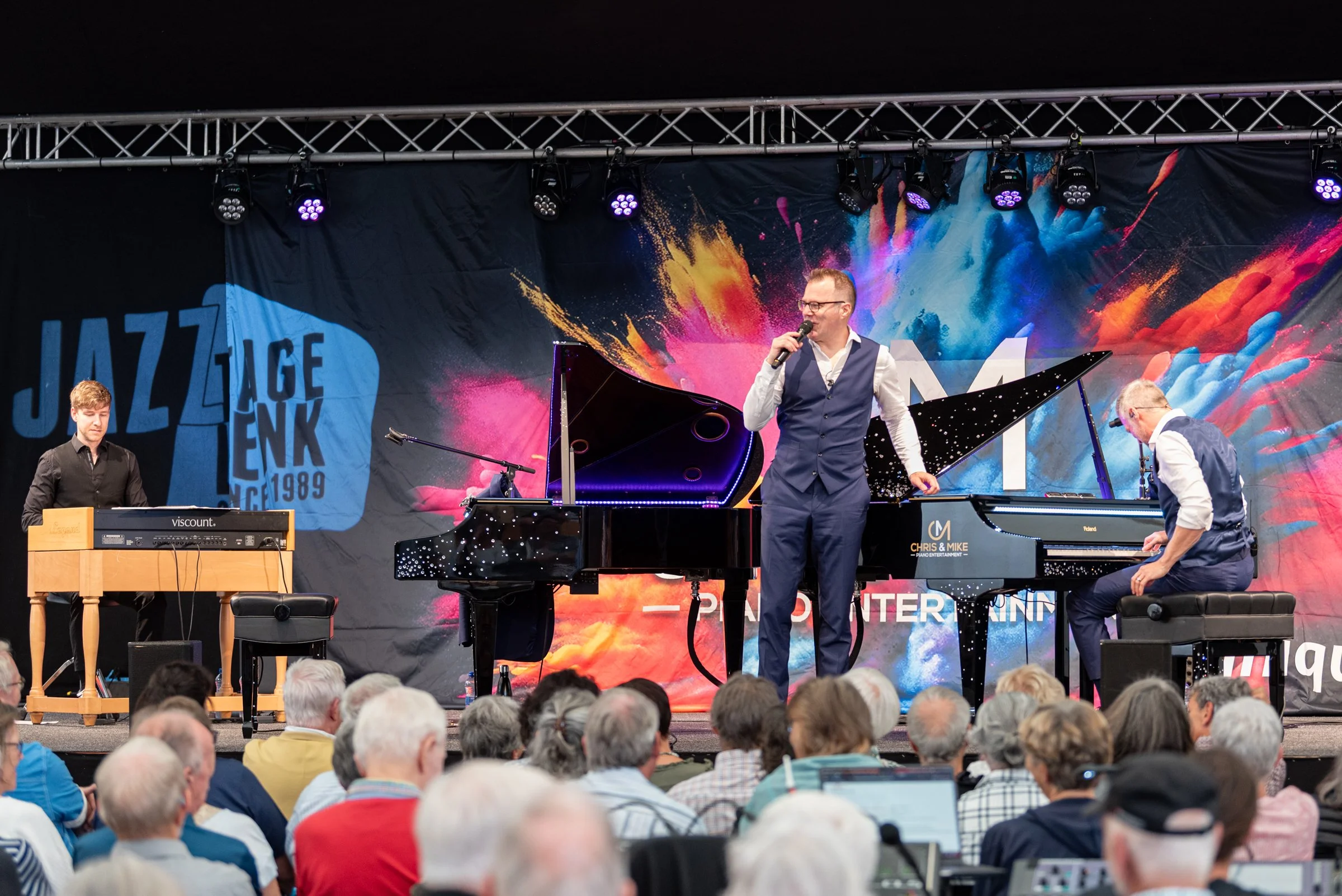 Jazzband auf Bühne während eines Konzerts vor Publikum, mit Schlagzeug, Klavier und Sänger, farbenfrohes Bühnenbild mit 'Jazz Jamboree 1989' Schriftzug.