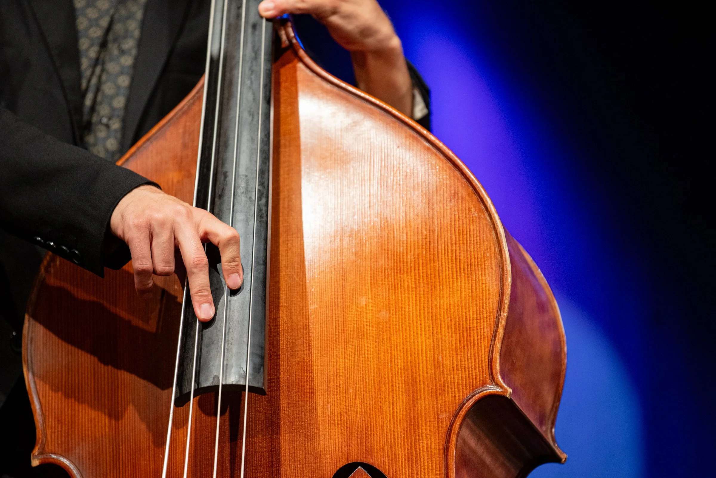Nahaufnahme eines klassischen Cello-Spielers, der auf einer Bühne spielt. Der Fokus liegt auf den Händen und dem Instrument, das aus Holz gefertigt ist, mit einer schwarzen Saitenhalterung.