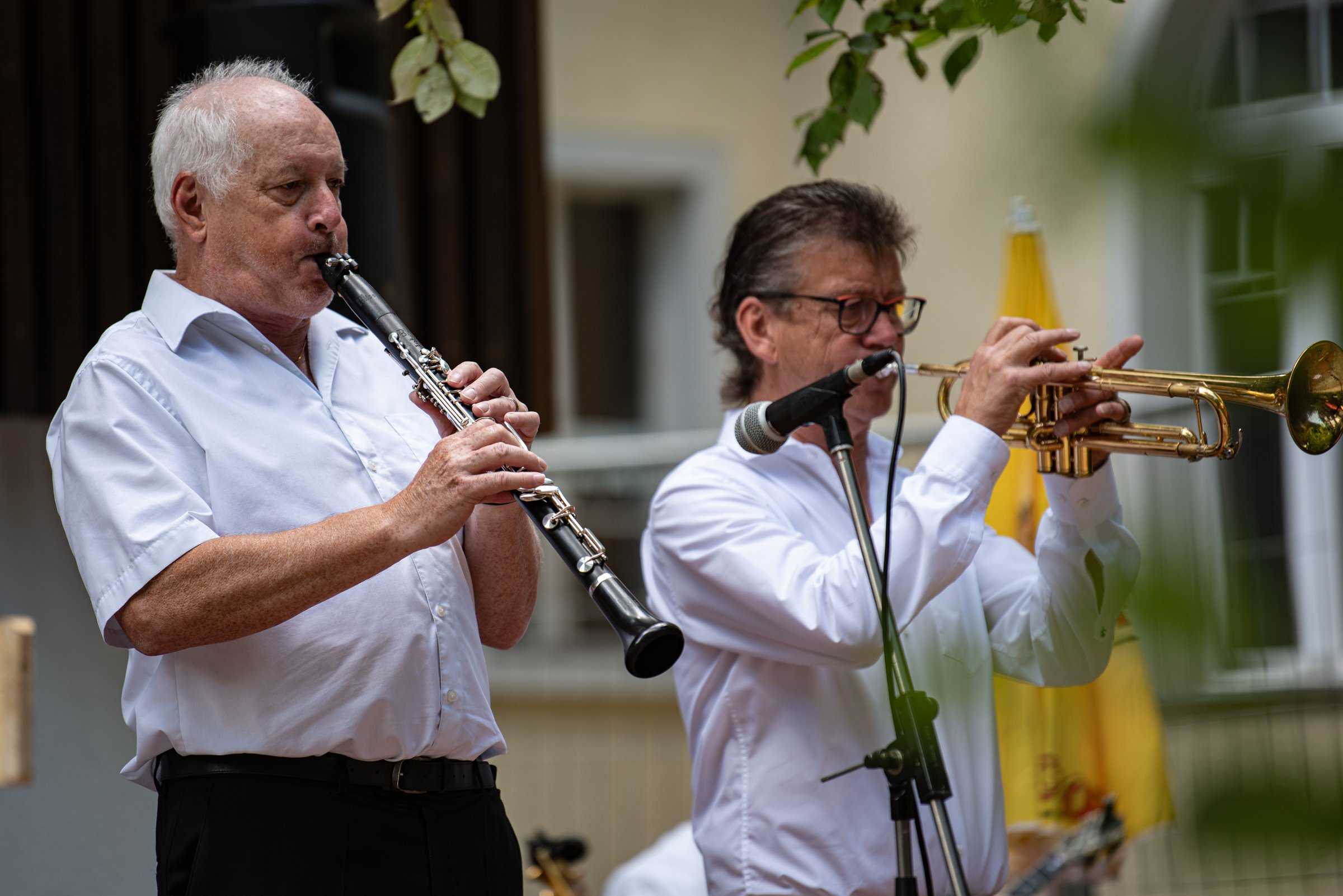 Zwei Männer spielen Musikinstrumente, der eine Klarinette, der andere Trompete, bei einer Outdoor-Veranstaltung.