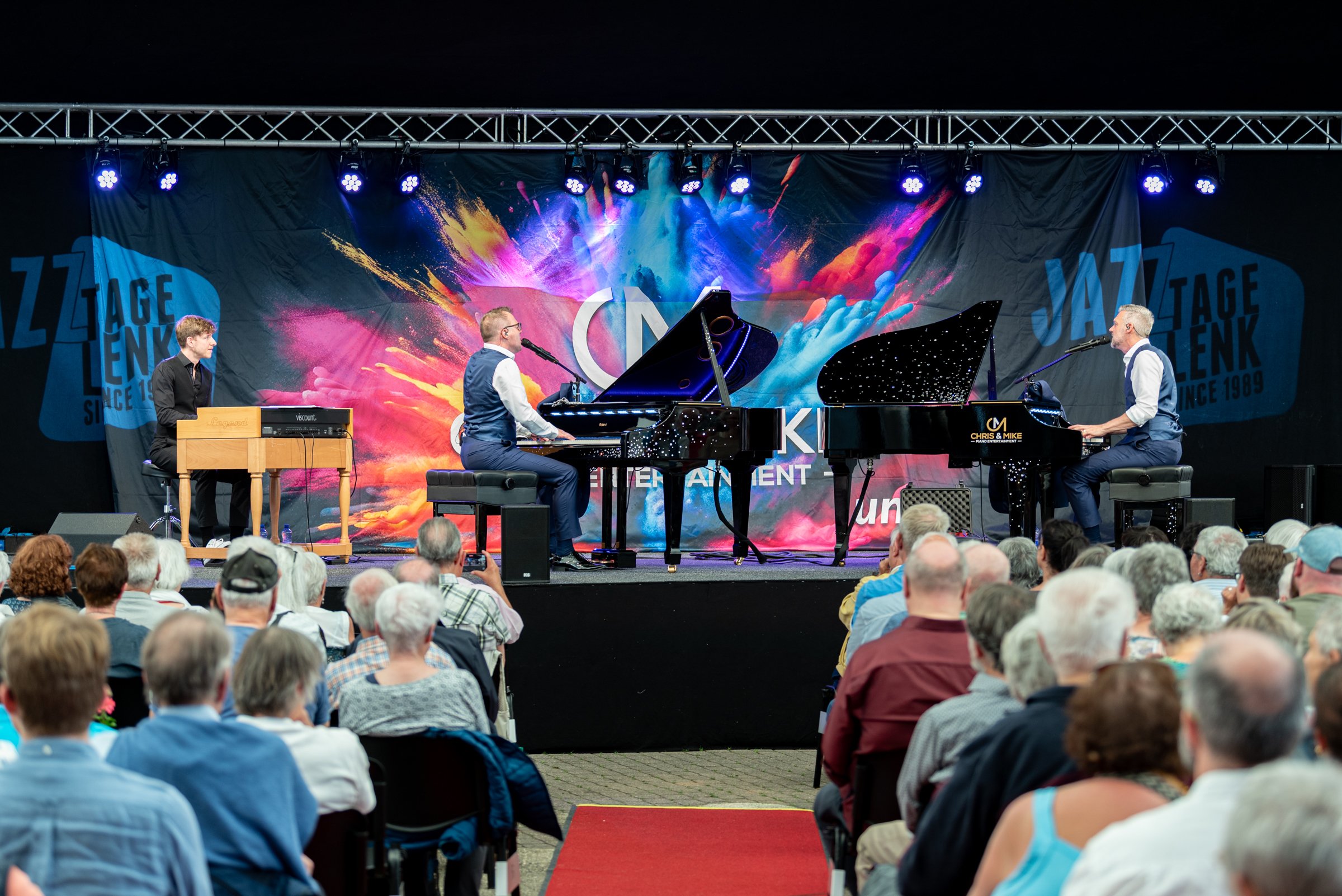 Musiker auf einer Bühne bei einem Konzert, drei Männer am Klavier, ein Mann begleitet auf einem kleineren Keyboard, vor einem Publikum an einem Abend. Ein bunter Hintergrund mit dem Text »JAZZ TAGE LÜNEBURG SINCE 1989«.