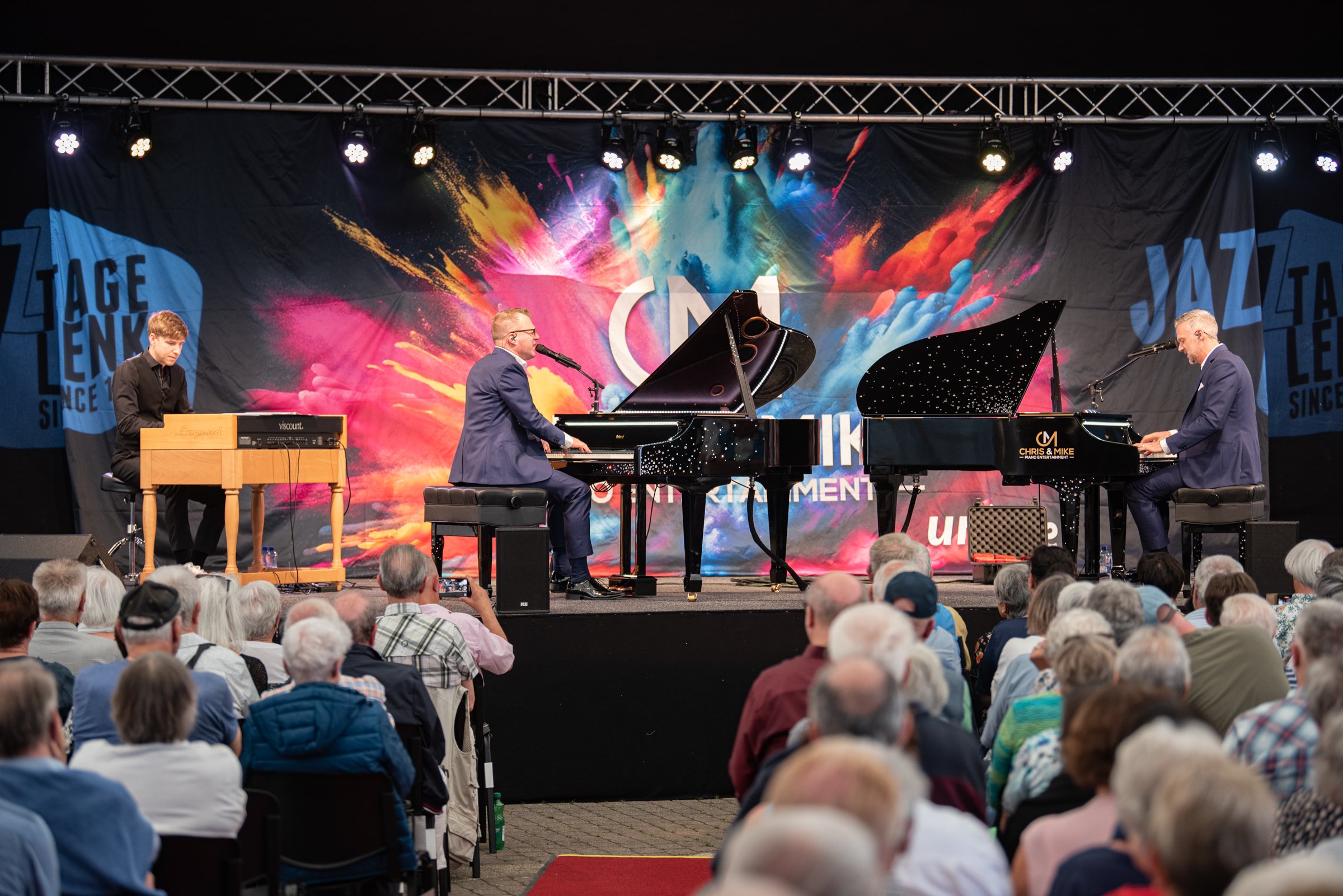 Drei Musiker spielen auf einer Bühne vor einem Publikum. Zwei spielen Klavier, einer spielt ein elektronisches Keyboard, im Hintergrund ein bunter, explosionartiger Hintergrund mit dem Text "JAZZ" und anderen Elementen.