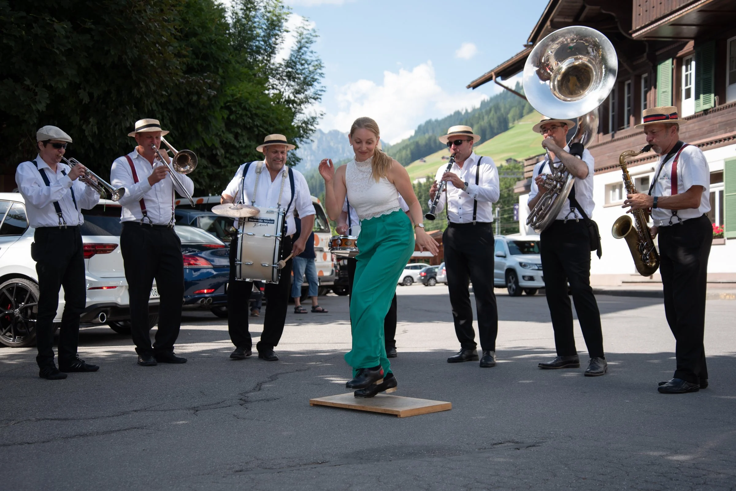 Eine Gruppe von Musikern spielt draußen, während eine Frau in einem weißen Top und grünen Hosen auf einem kleinen Holzblock tanzt.