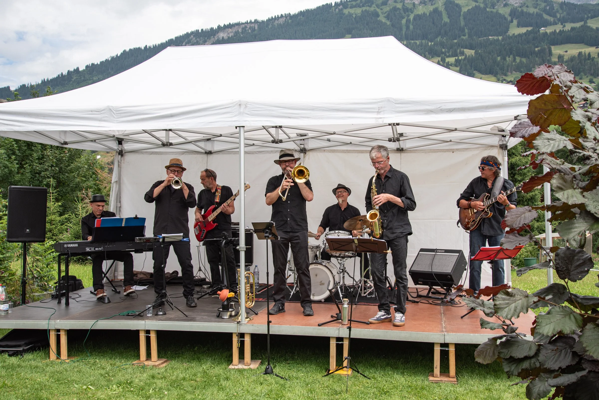 Eine Band mit sechs Mitgliedern spielt auf einer kleinen Bühne im Freien, umgeben von grüner Natur und Bergen im Hintergrund. Sie spielen verschiedene Instrumente, darunter Klavier, Trompete, Bass, Schlagzeug, Saxophon und Gitarre.