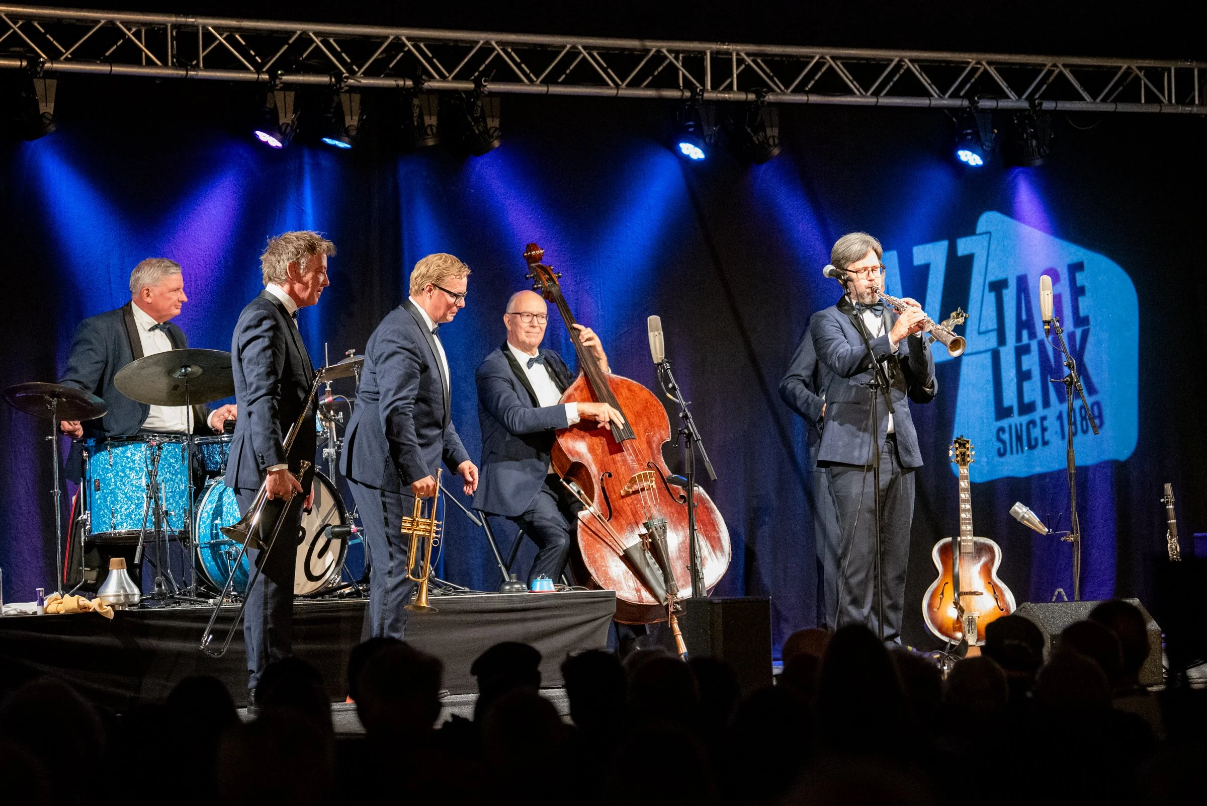 Eine Band mit sechs männlichen Musikern in formeller Kleidung spielt auf einer Bühne. Zwei Gitarren, ein Kontrabass, ein Schlagzeug und ein Saxophon sind sichtbar, alle vor einem blauen Vorhang mit einem Logo.