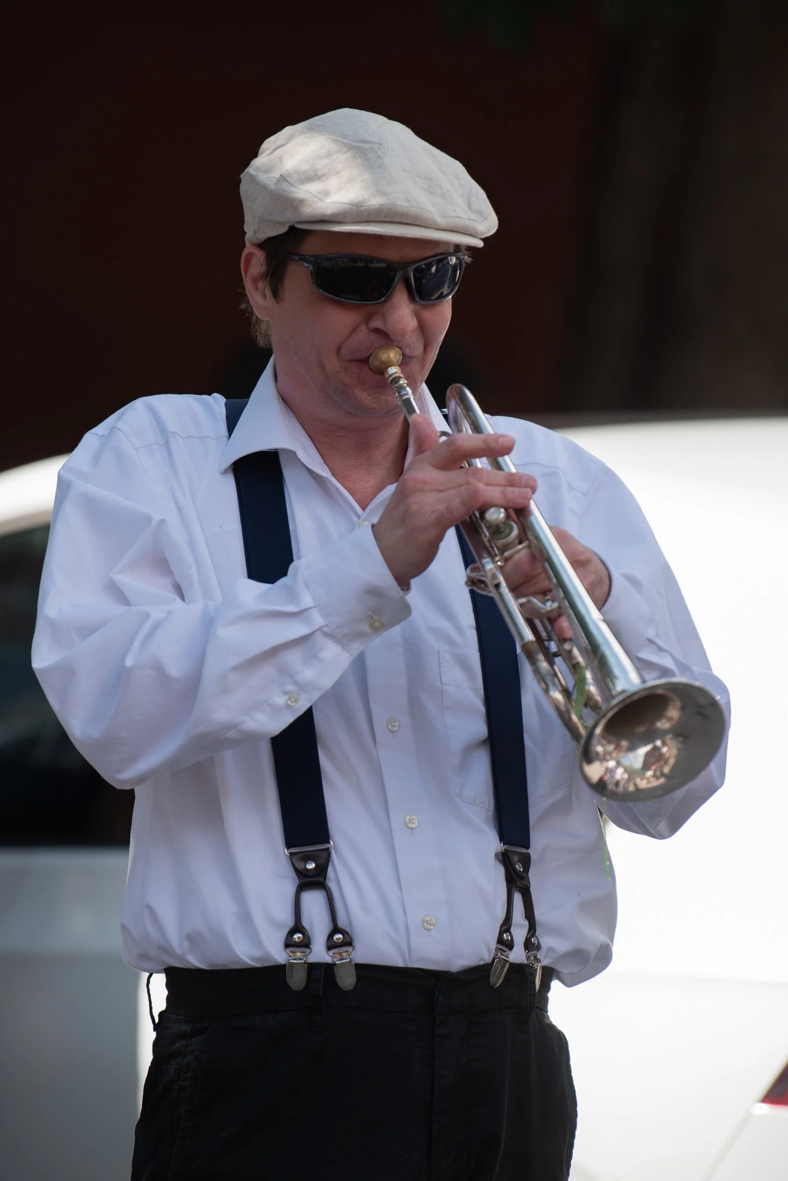 Mann spielt Trompete, trägt weißen Hemd, schwarze Sonnenbrille, Schal, Hut und Hosenträger.