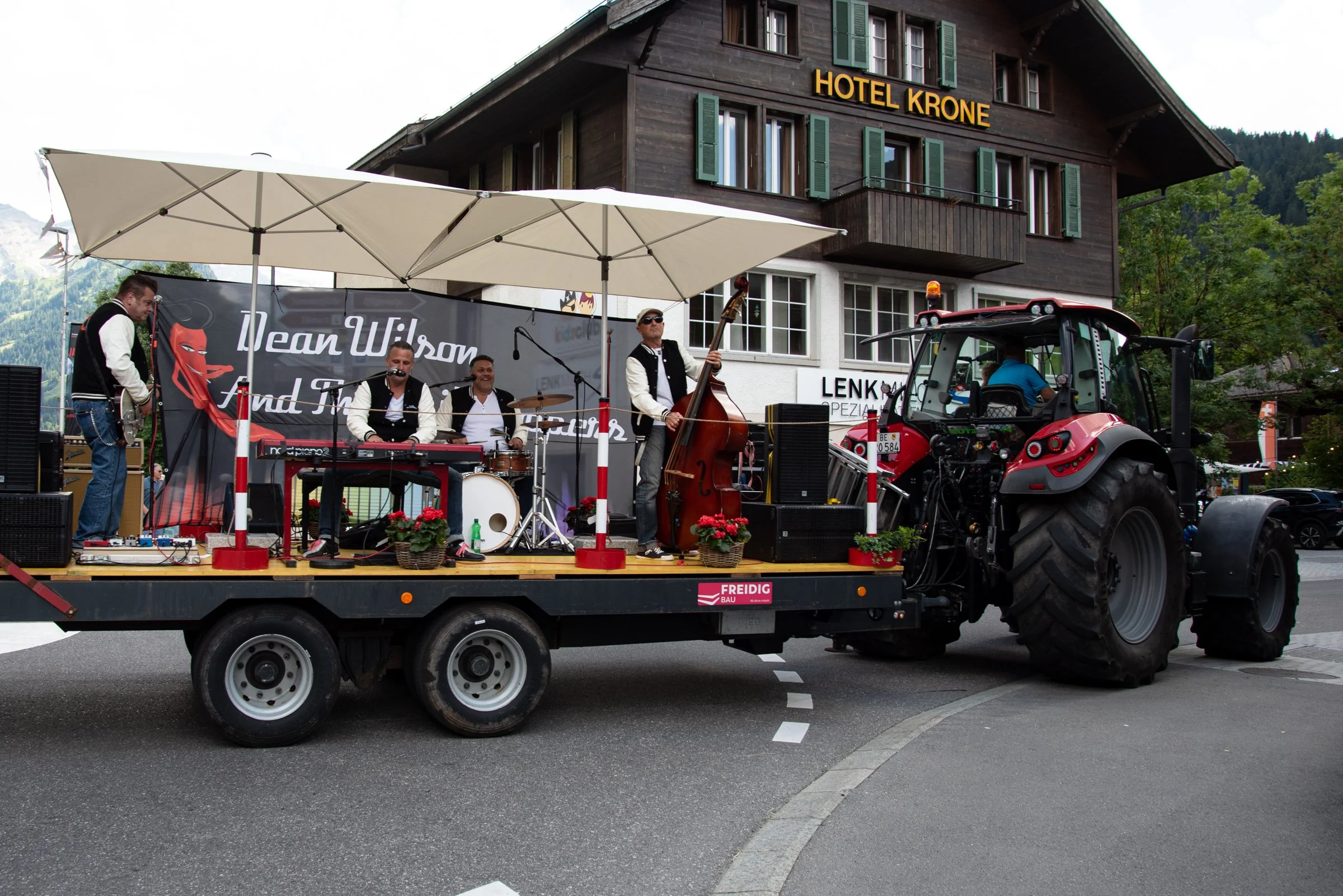 Musiker spielen auf einer fahrenden Plattform, die an einen Traktor angebunden ist, vor einem Hotel mit Holzarchitektur, in der Schweiz, mit einem Berg im Hintergrund.