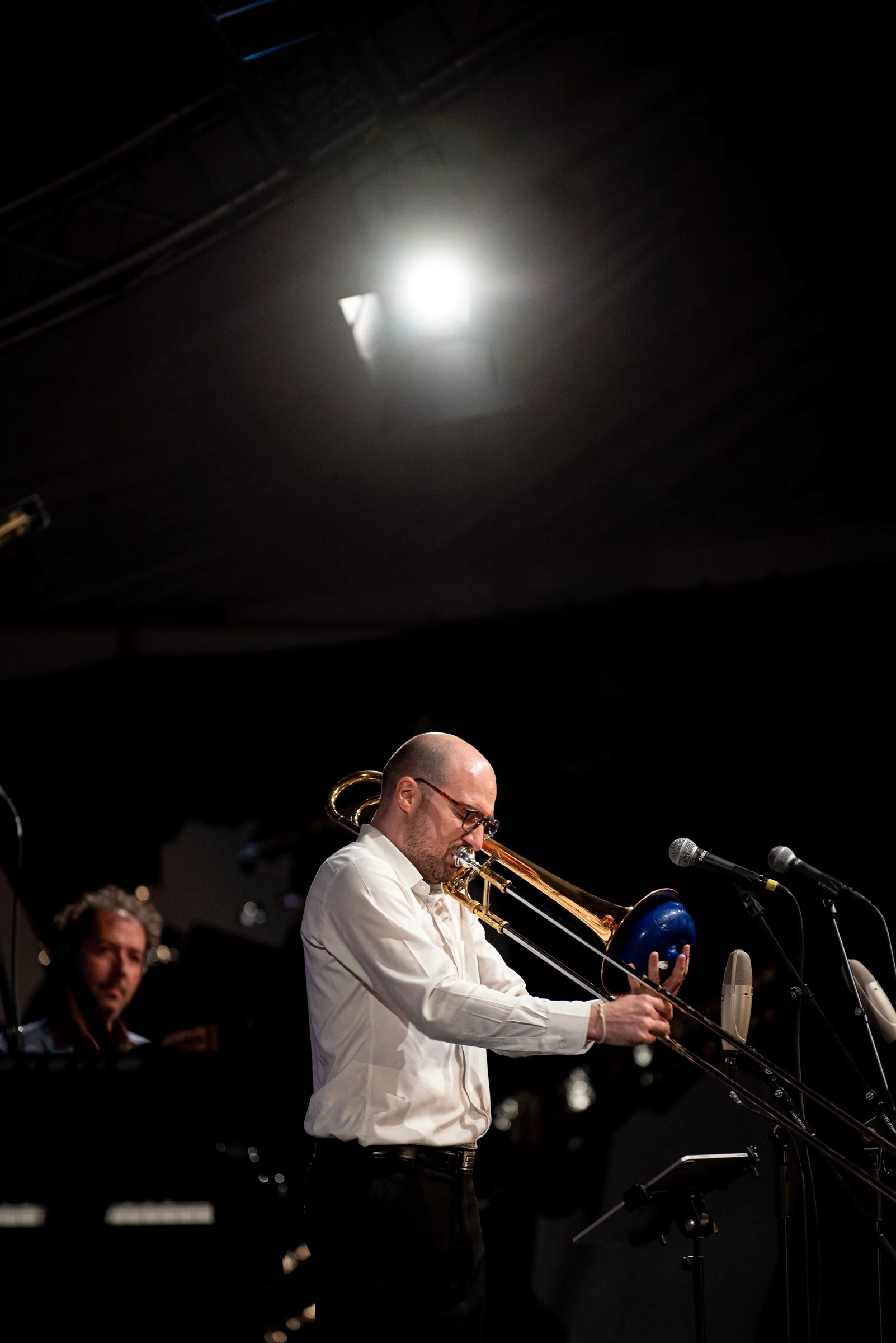 Ein Mann spielt Trompete auf einer Bühne, im Hintergrund ein Pianospieler, mit Mikrofonen und dunklem Bühnenhintergrund.