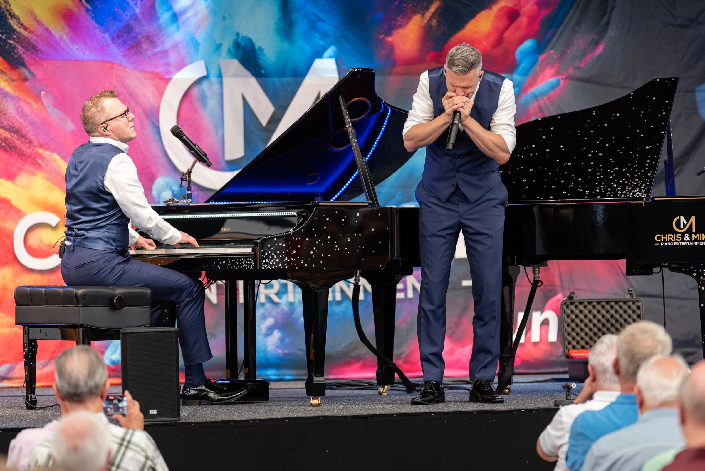 Zwei Musiker bei einer Bühnenperformance, einer spielt Klavier, der andere singt in ein Mikrofon, im Hintergrund ein bunten, kreatives Bühnenbild