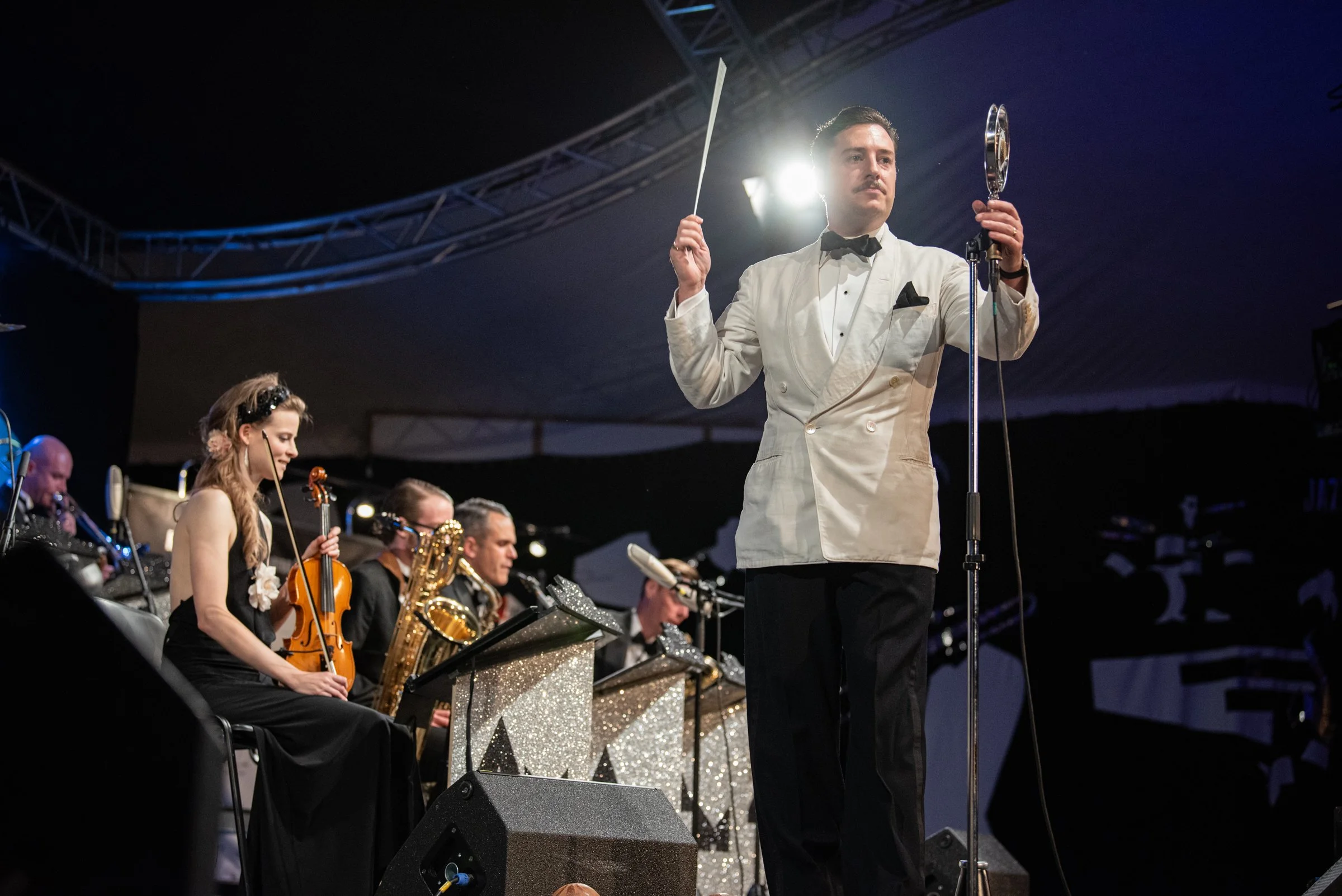 Ein Musiker im weißen Frack dirigiert ein Orchester bei einem Konzert auf einer Bühne. Im Hintergrund sitzen weitere Musiker mit verschiedenen Instrumenten.