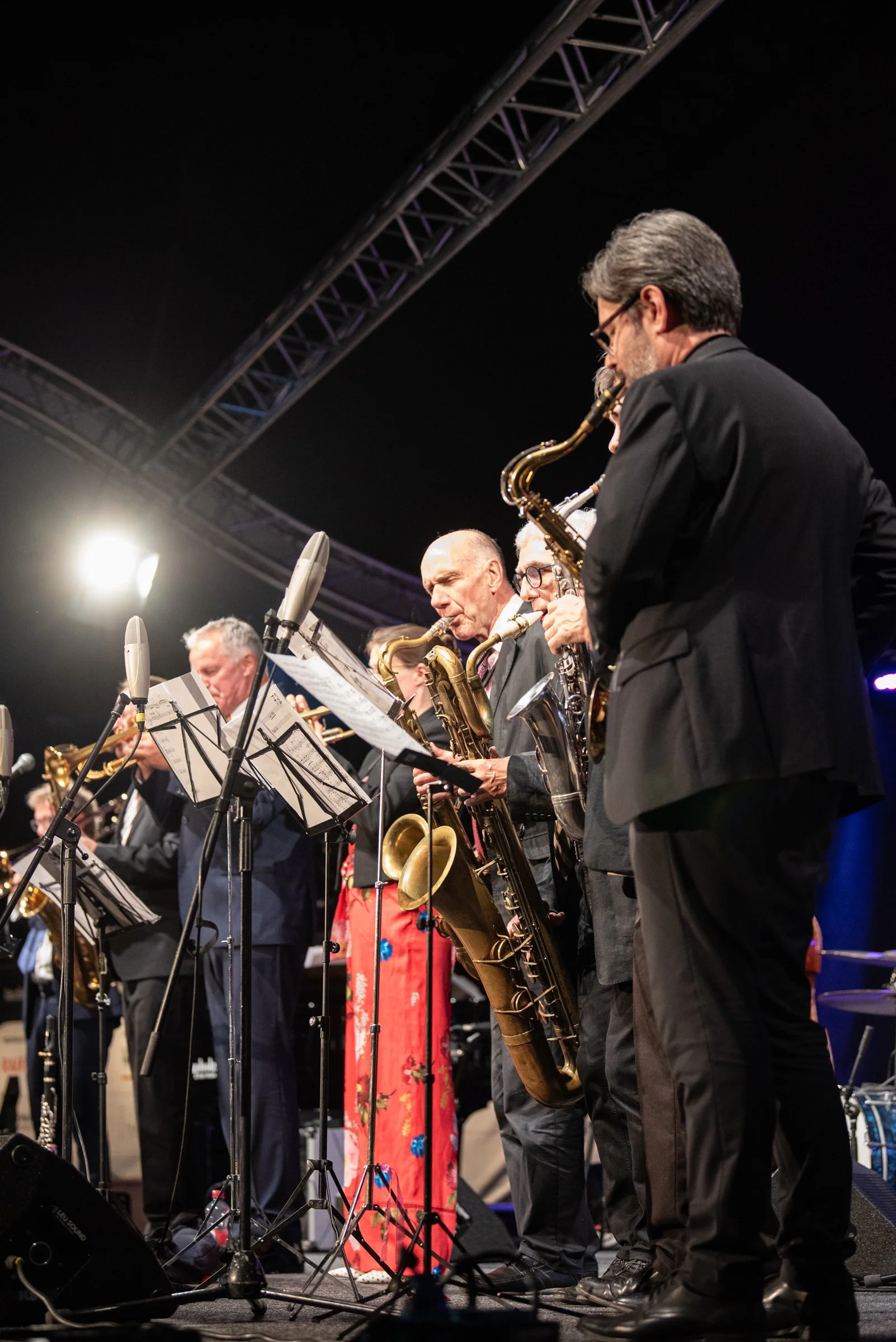 Musiker auf der Bühne spielen Saxophon, Mikrofone und Notenständer im Einsatz, dunkler Hintergrund, Bühnenbeleuchtung.