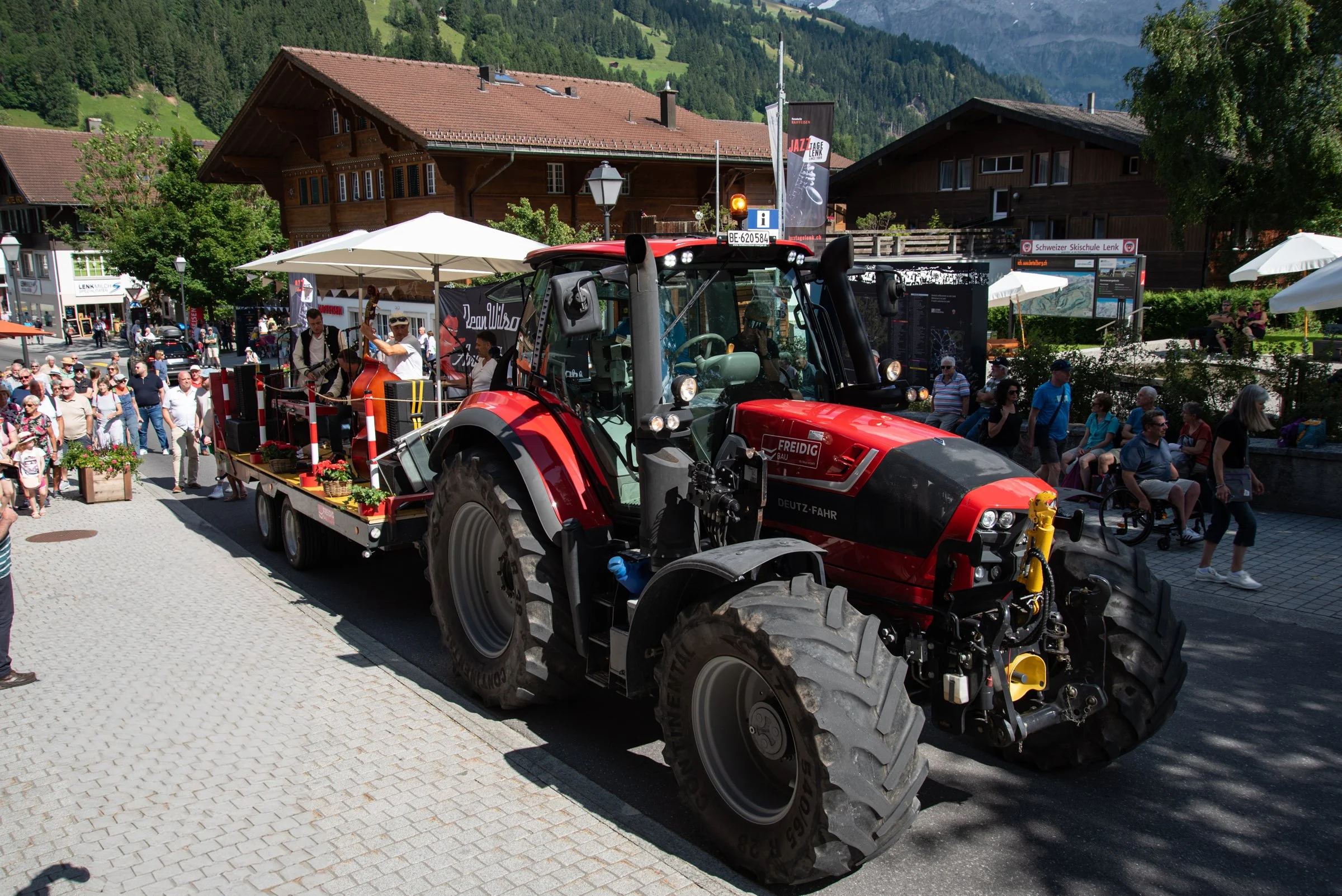 Ein roter Traktor mit einem Anhänger voller Blumen steht auf einer Straße in einer belebten Ortslage, umgeben von Menschen bei einem Stadtfest oder Markt in einer ländlichen Gegend, im Hintergrund sind traditionelle Holzchalets und grüne Hügel zu seh