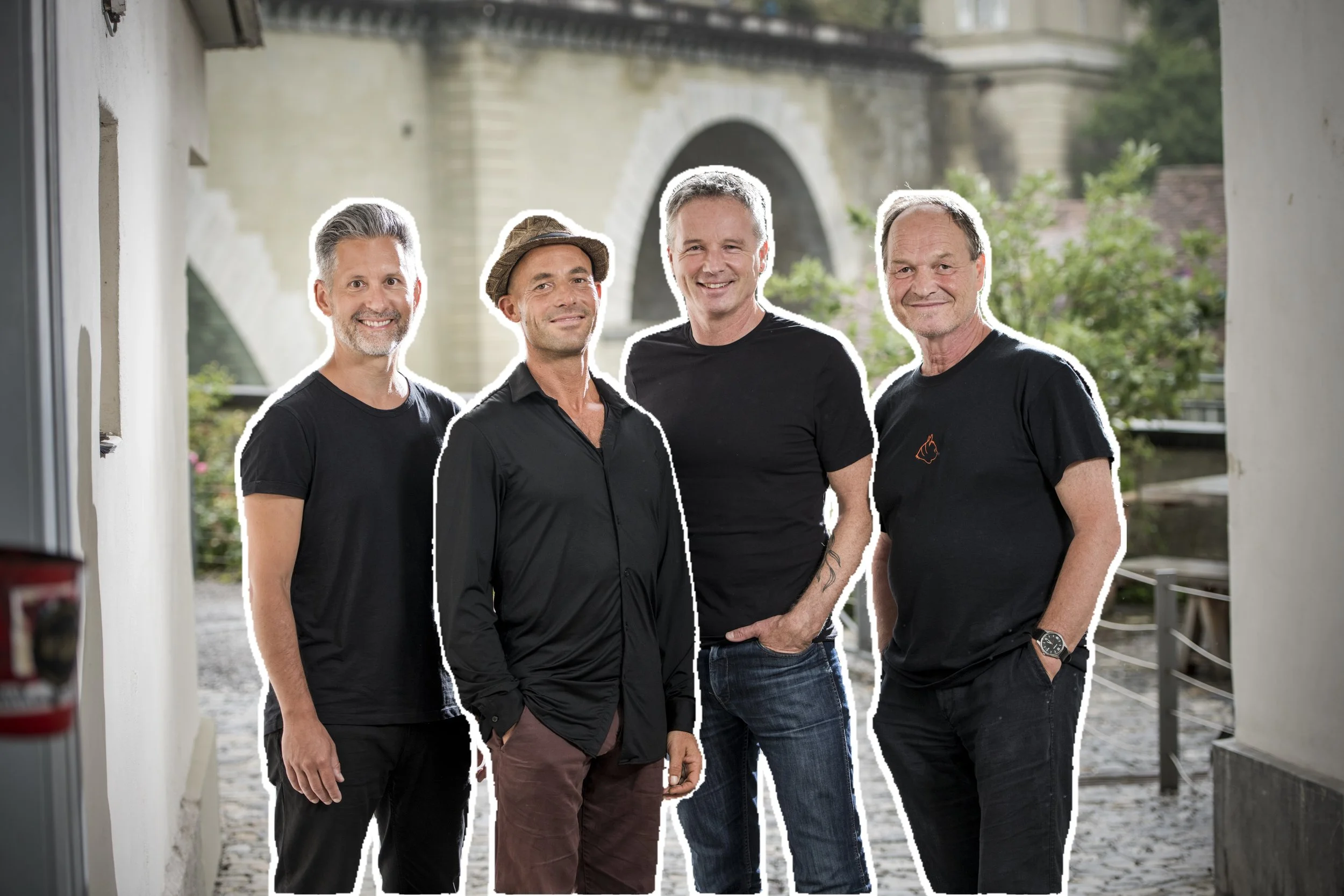 Vier Männer stehen nebeneinander im Freien vor einer alten Brücke, lächeln in die Kamera, tragen schwarze Kleidung, einige mit Hand in Tasche, eines trägt eine Armbanduhr, im Hintergrund sind Bäume und eine historische Brücke sichtbar.