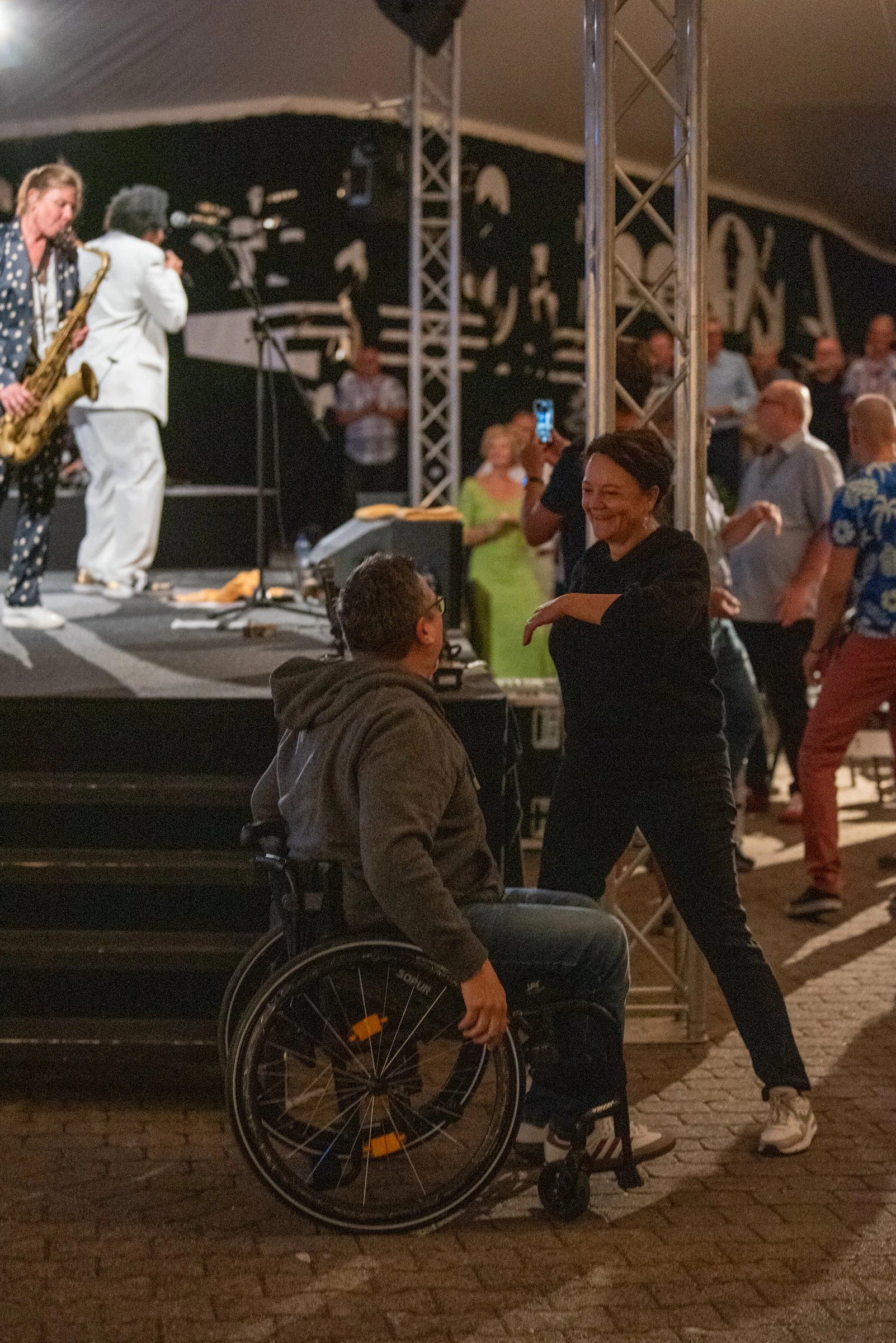 Menschen tanzen und ein Mann im Rollstuhl sitzt vor einer Bühne, während eine Frau ihn anspricht. Hinter ihnen spielen Musiker auf der Bühne.