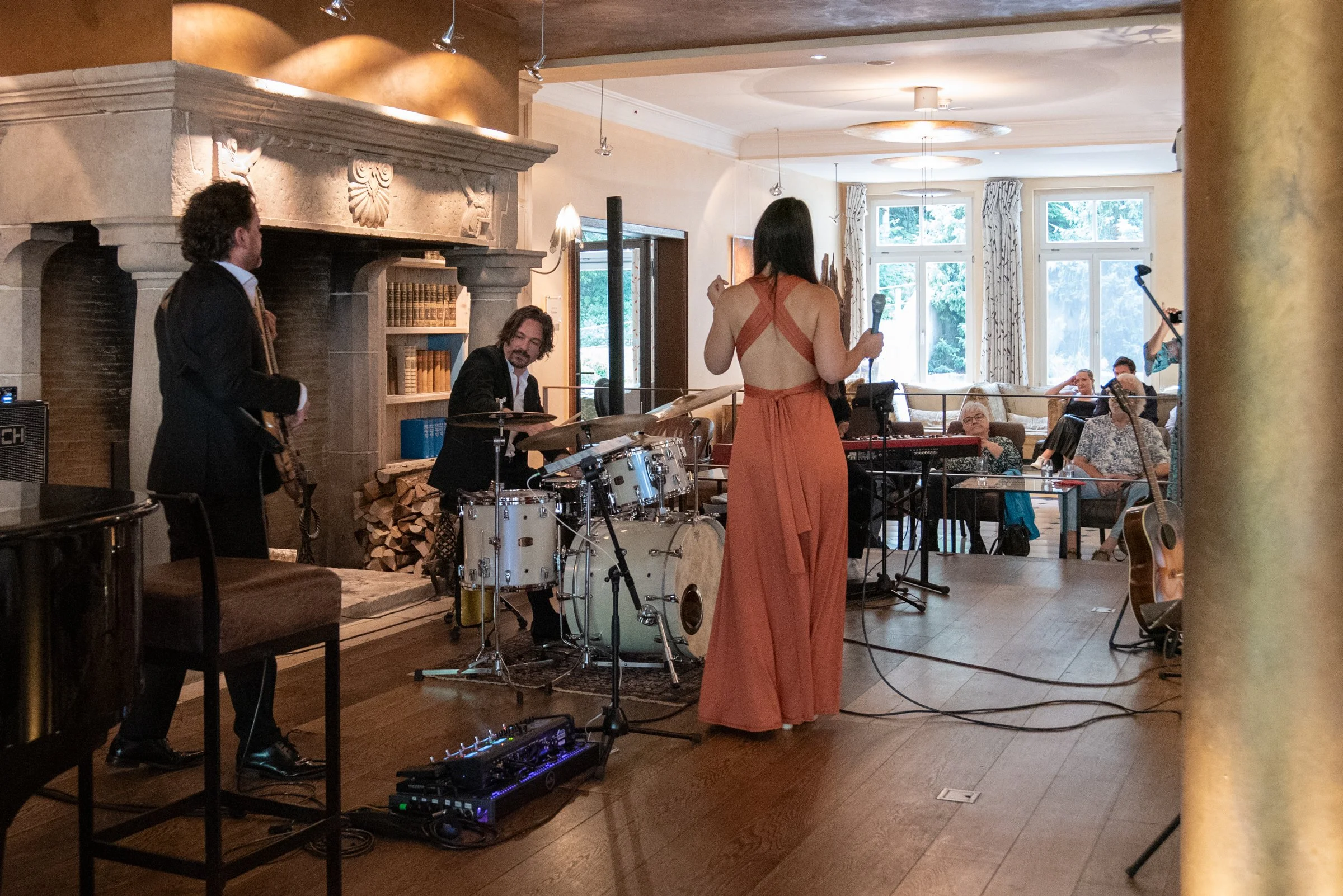 Eine Live-Jazzband spielt in einem gemütlichen Saal mit einer Frau, die einen schwarzen Mikrofon hält, während die Musiker mit Schlagzeug, Gitarre und Saxophon spielen, während das Publikum im Hintergrund sitzt.